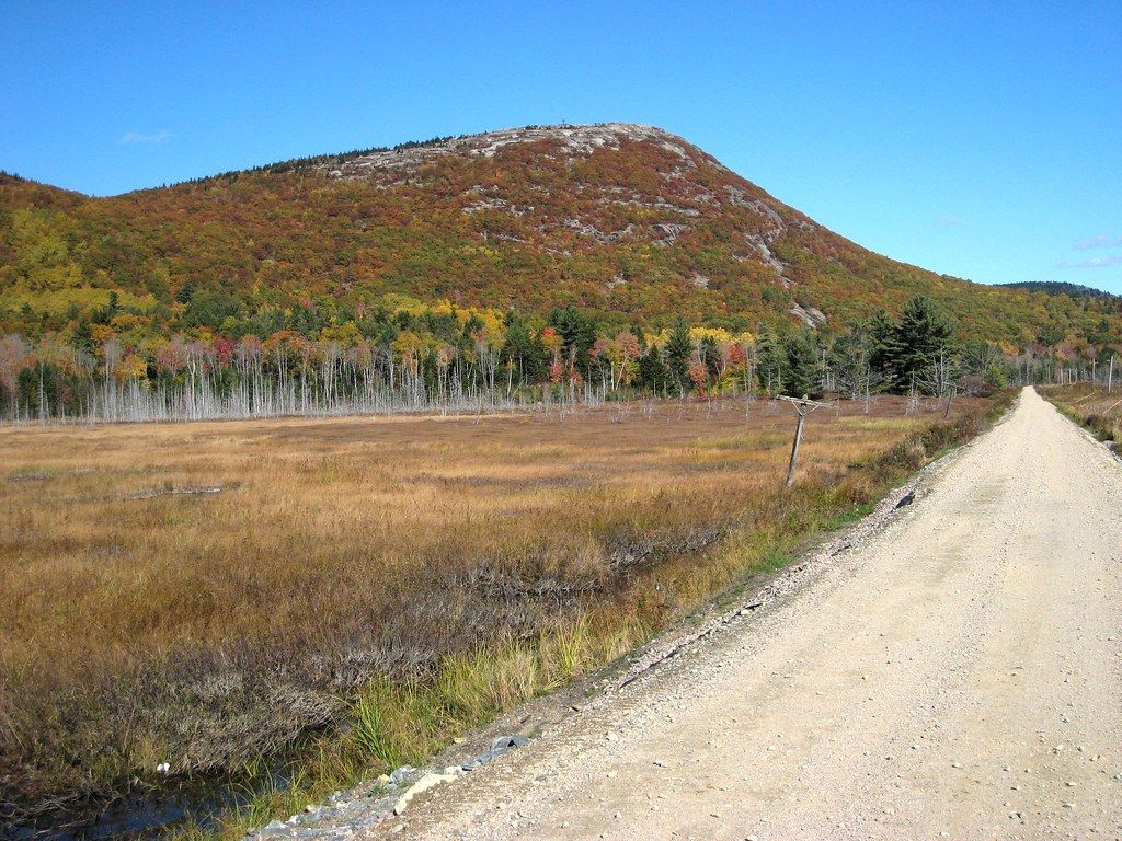 A dirt road leads past a field towards a mountain with autumn trees. - Down East Sunrise Trail