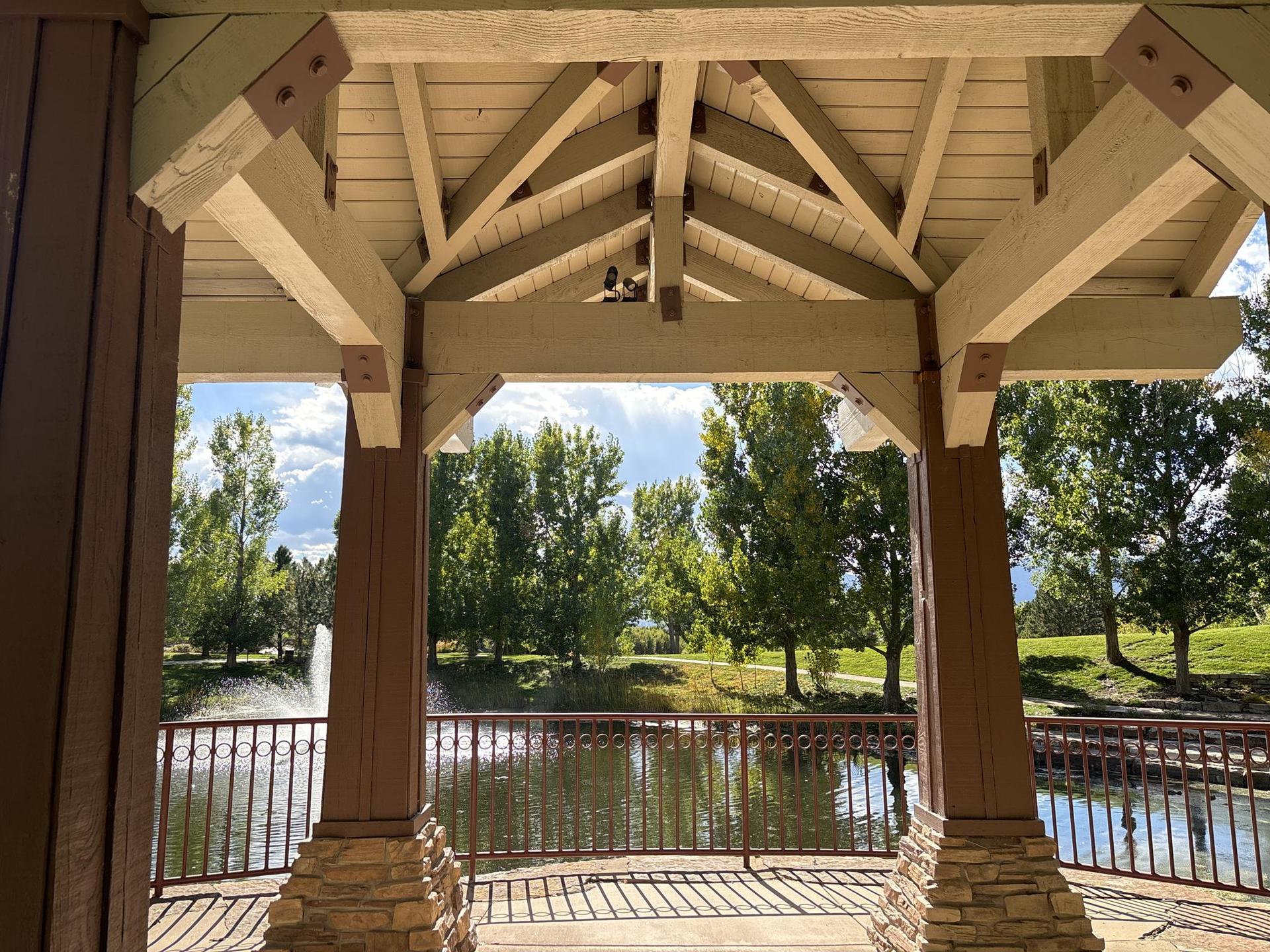 View from inside a wooden gazebo overlooking a pond with a fountain and trees.