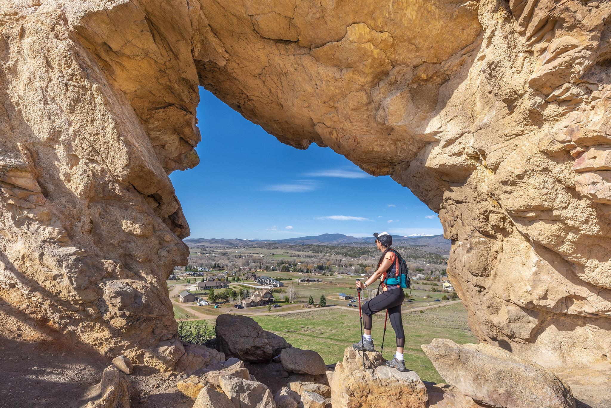 Hiker with poles stands in a rock arch overlooking a valley.