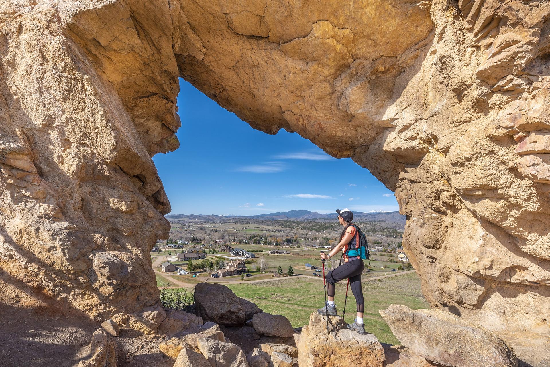 Hiker with poles stands in a rock arch overlooking a valley.