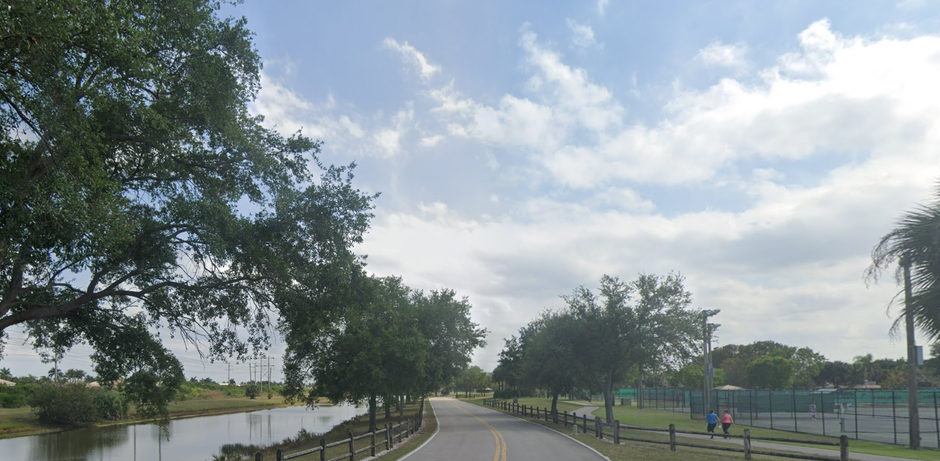 A paved road runs beside a lake and tennis courts under a cloudy sky.