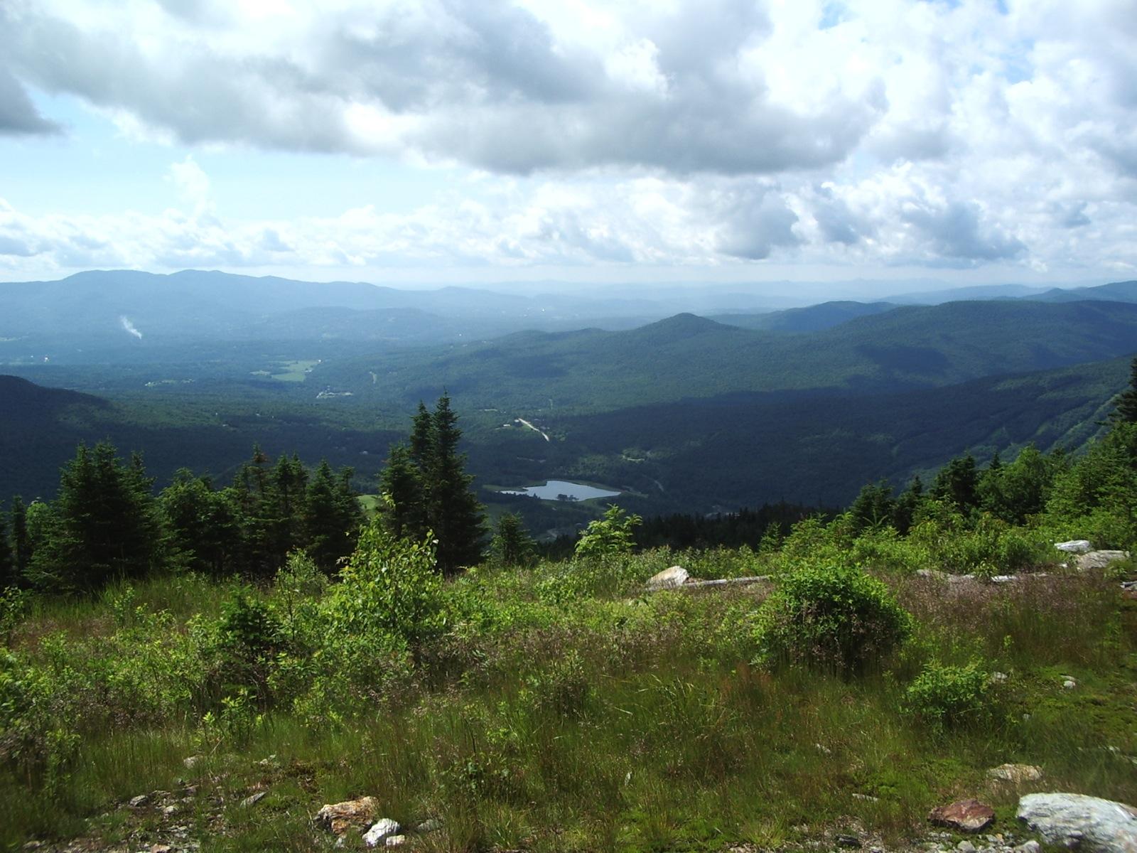 Expansive mountain vista with a lake under a bright, cloudy sky.