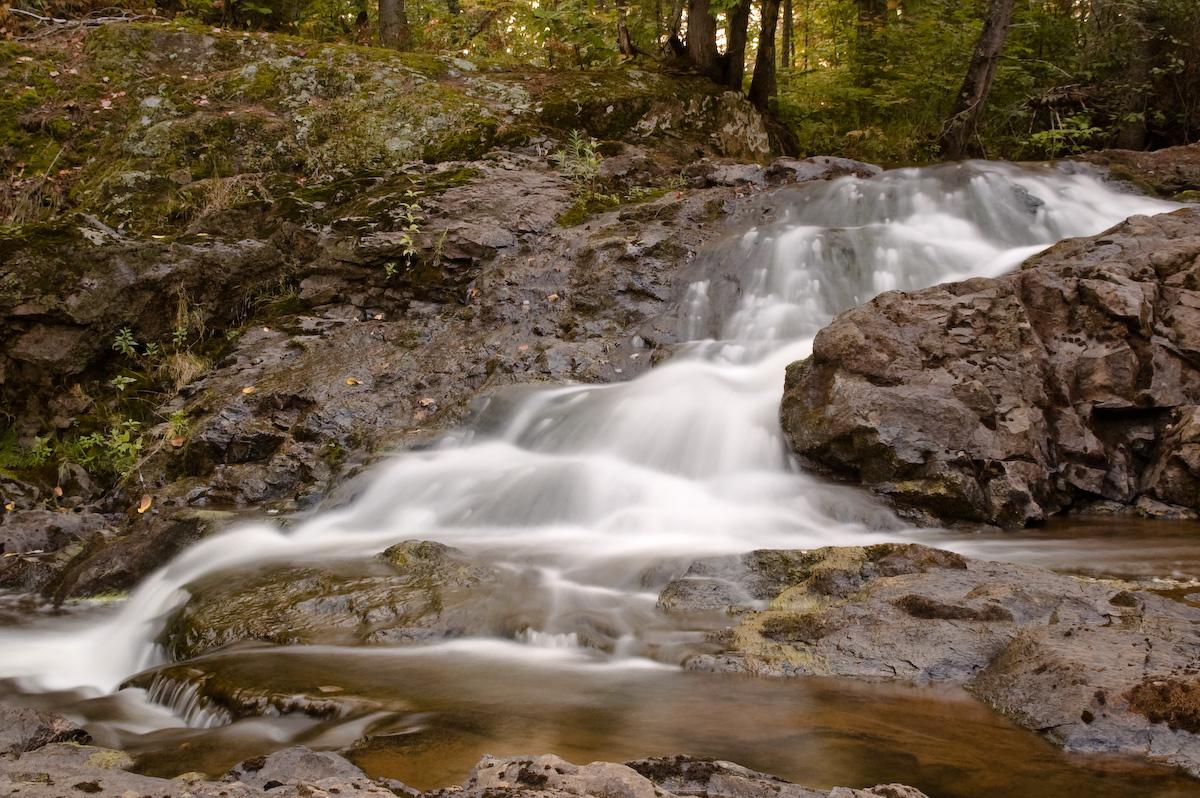 A blurred waterfall cascades over dark rocks in a lush, natural forest.
