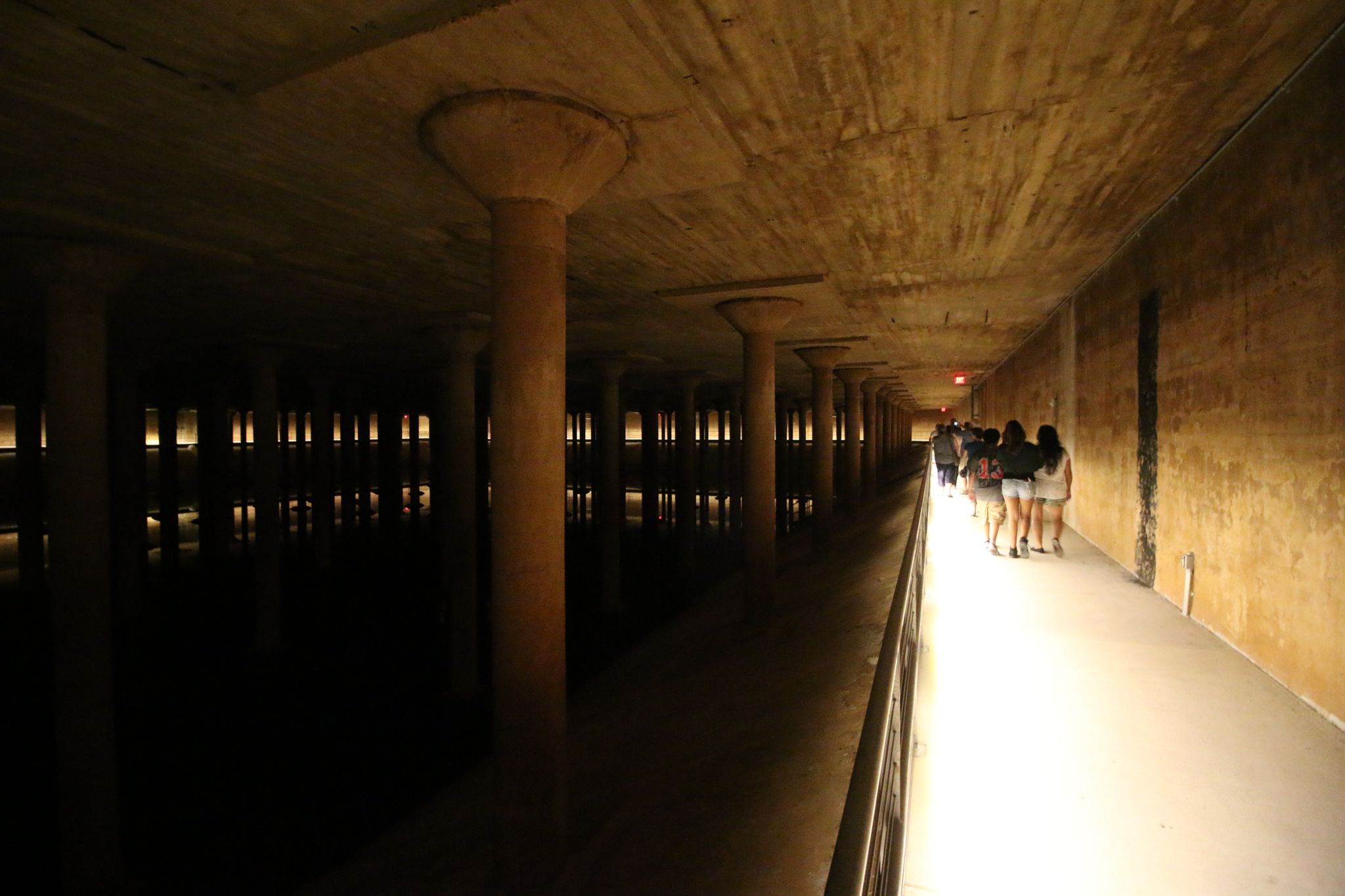 People walk along a dimly lit concrete pathway in a vast underground space. - Buffalo Bayou Loop Trail