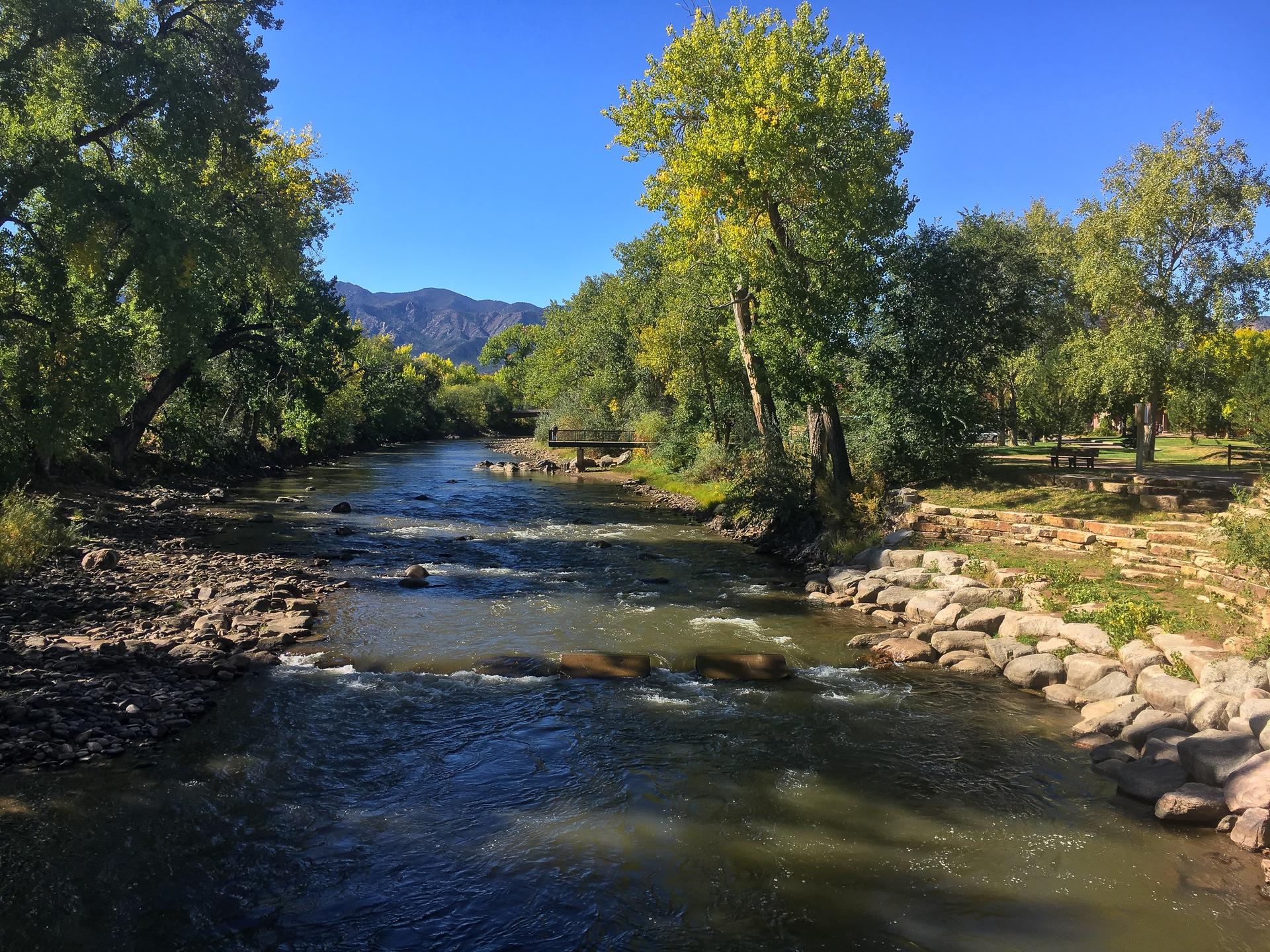 Sunny river scene with rocky banks, lush trees, and distant mountains under blue sky.