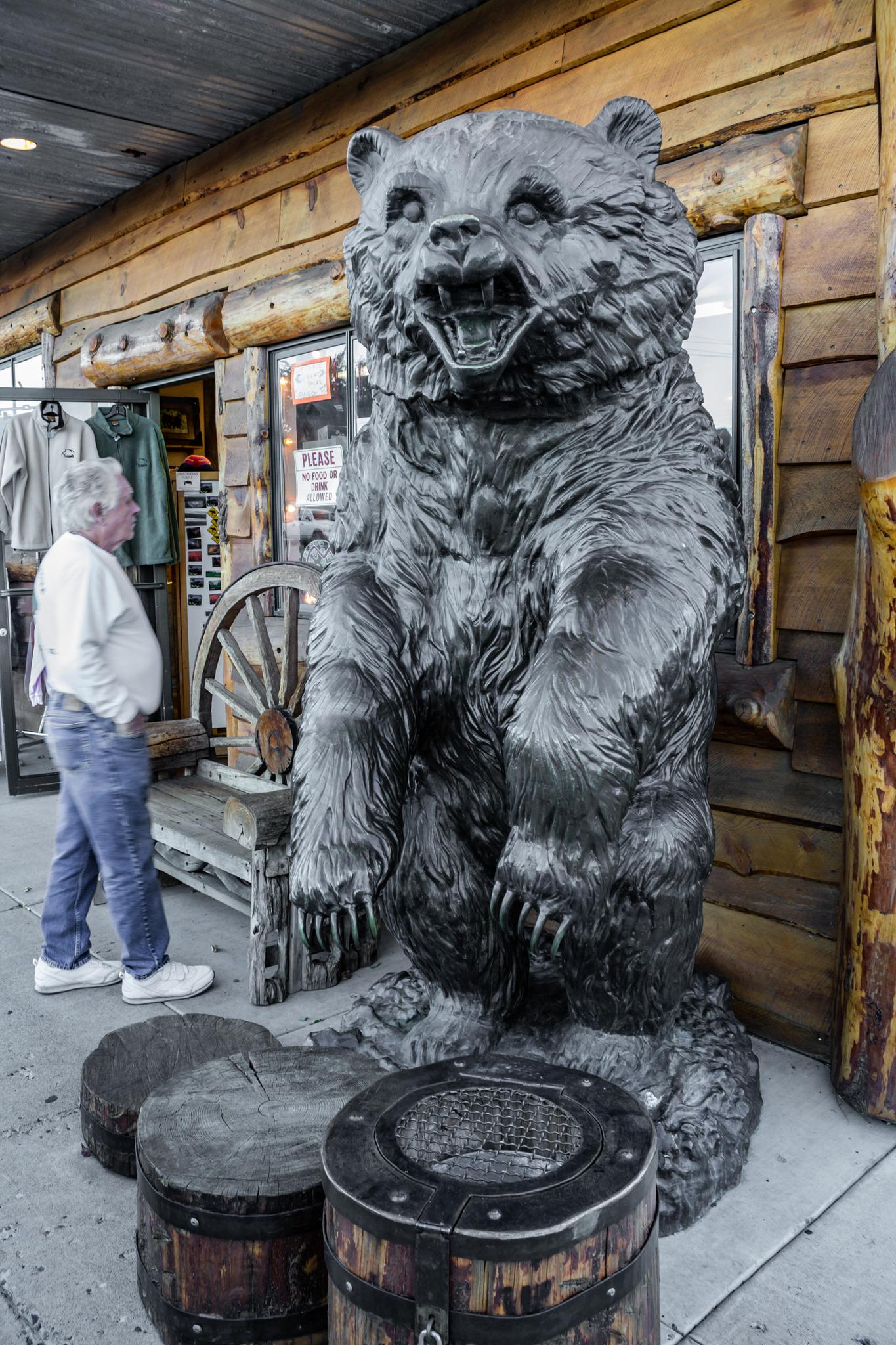 A man stands next to a large, dark bear statue outside a wooden building.