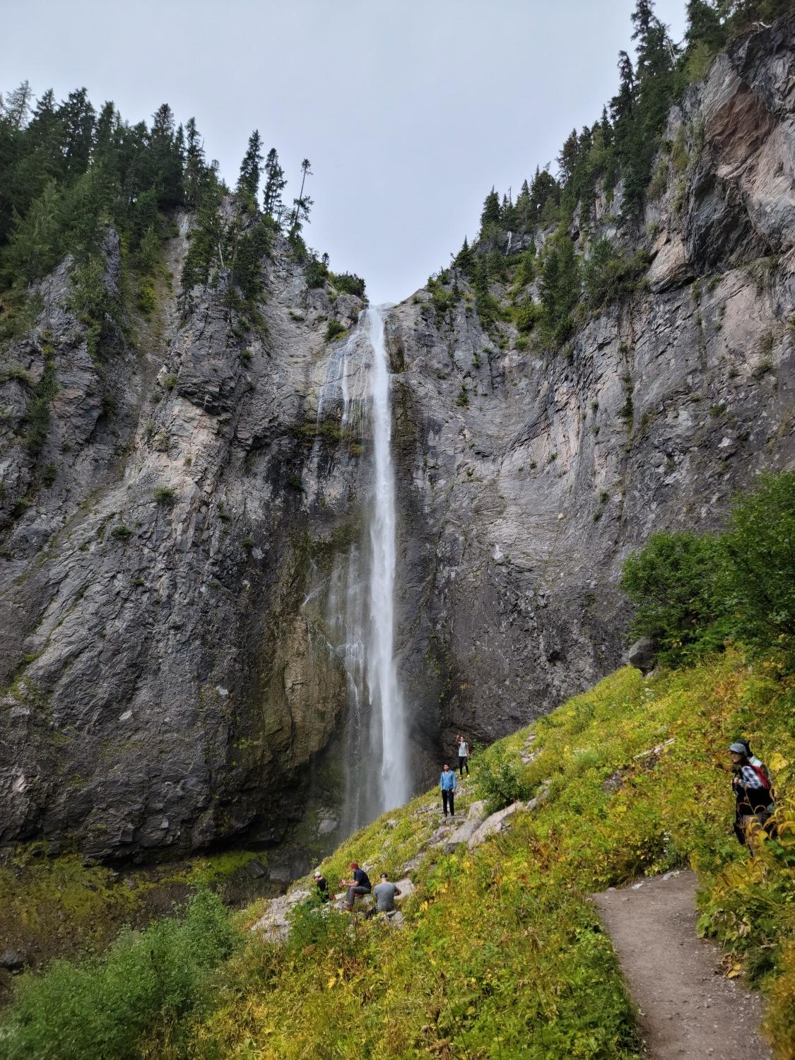 Tall waterfall cascading down a cliff with people on a grassy slope.
