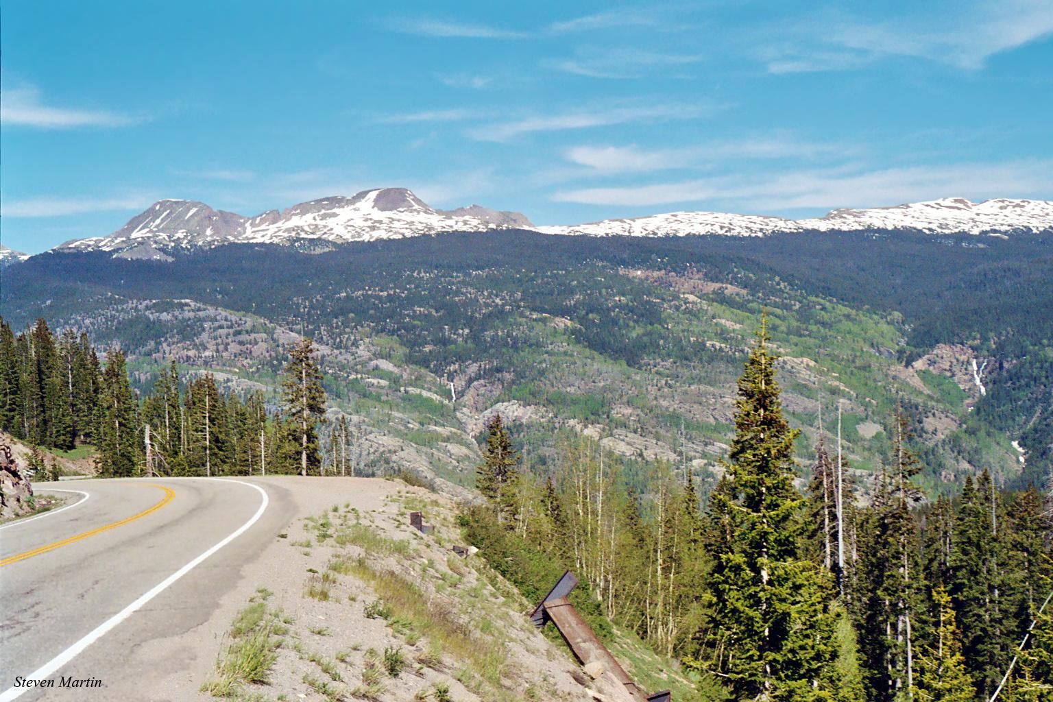 Sunny mountain road curves towards distant snow-capped peaks and forested valleys.