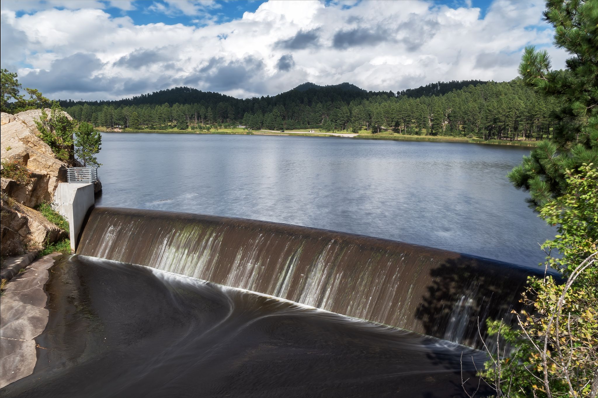 A dam with water flowing over it, leading to a calm lake and forested mountains. - Custer loop via Needles Highway