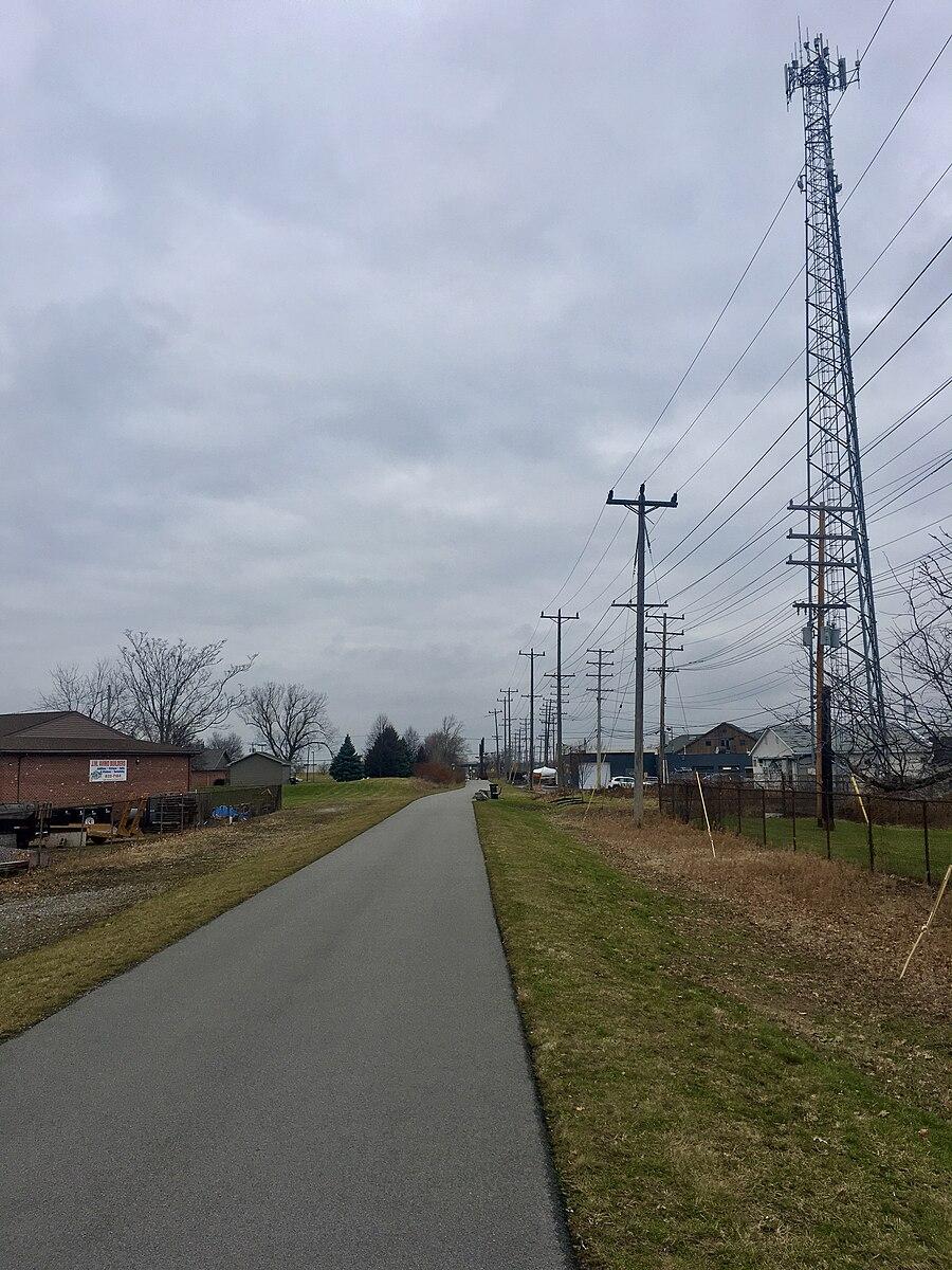 Paved path alongside utility poles and a cell tower under an overcast sky.
