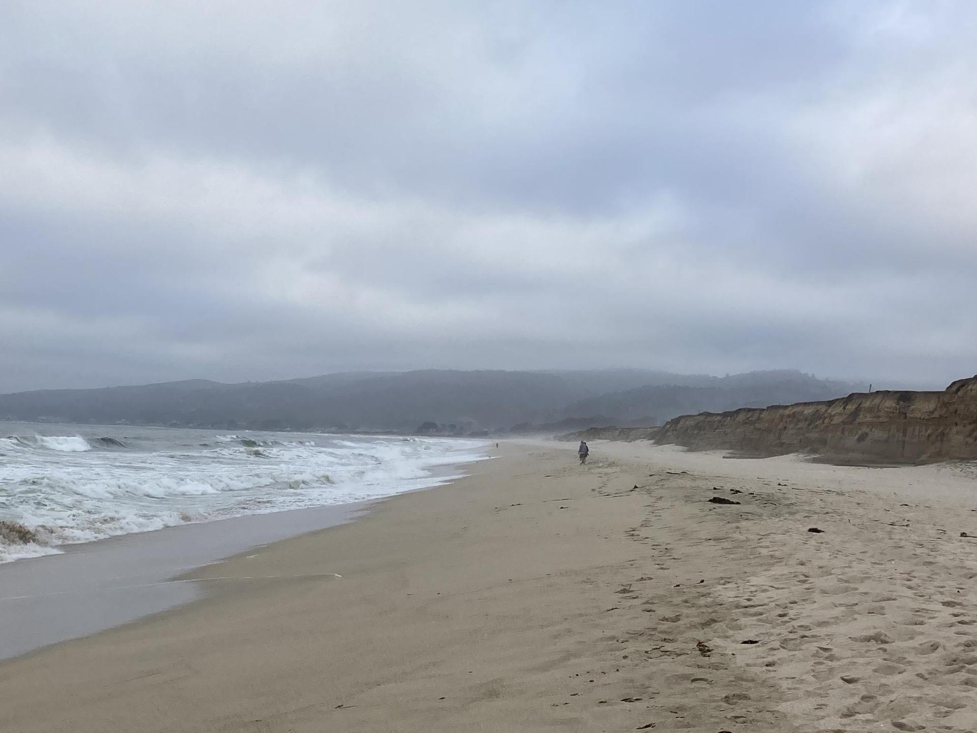 Wide sandy beach with crashing waves, eroded cliffs, and a lone walker.