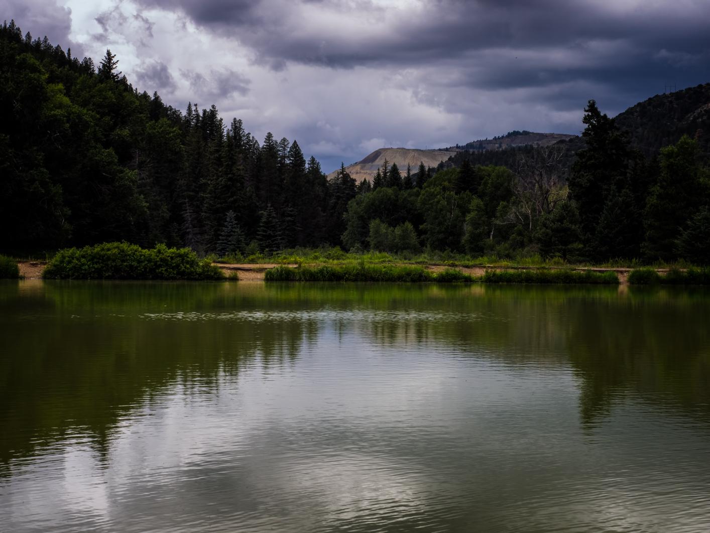 A dark, moody lake reflects a forested mountain under cloudy skies.