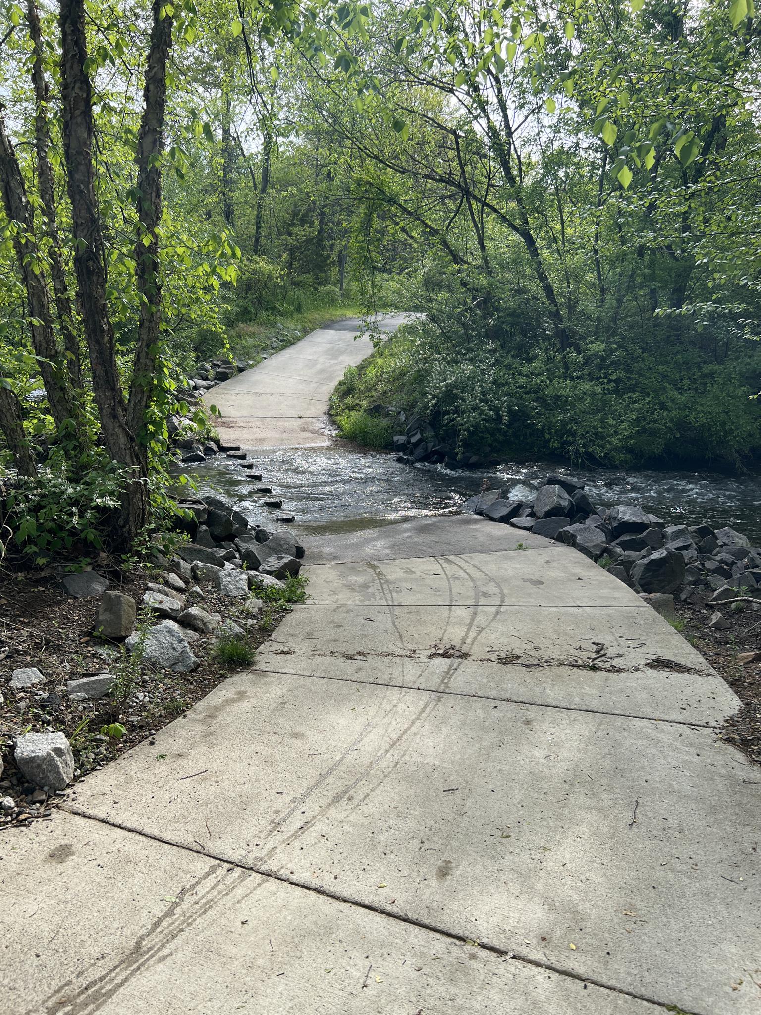Concrete path descends into a flowing stream surrounded by green trees.