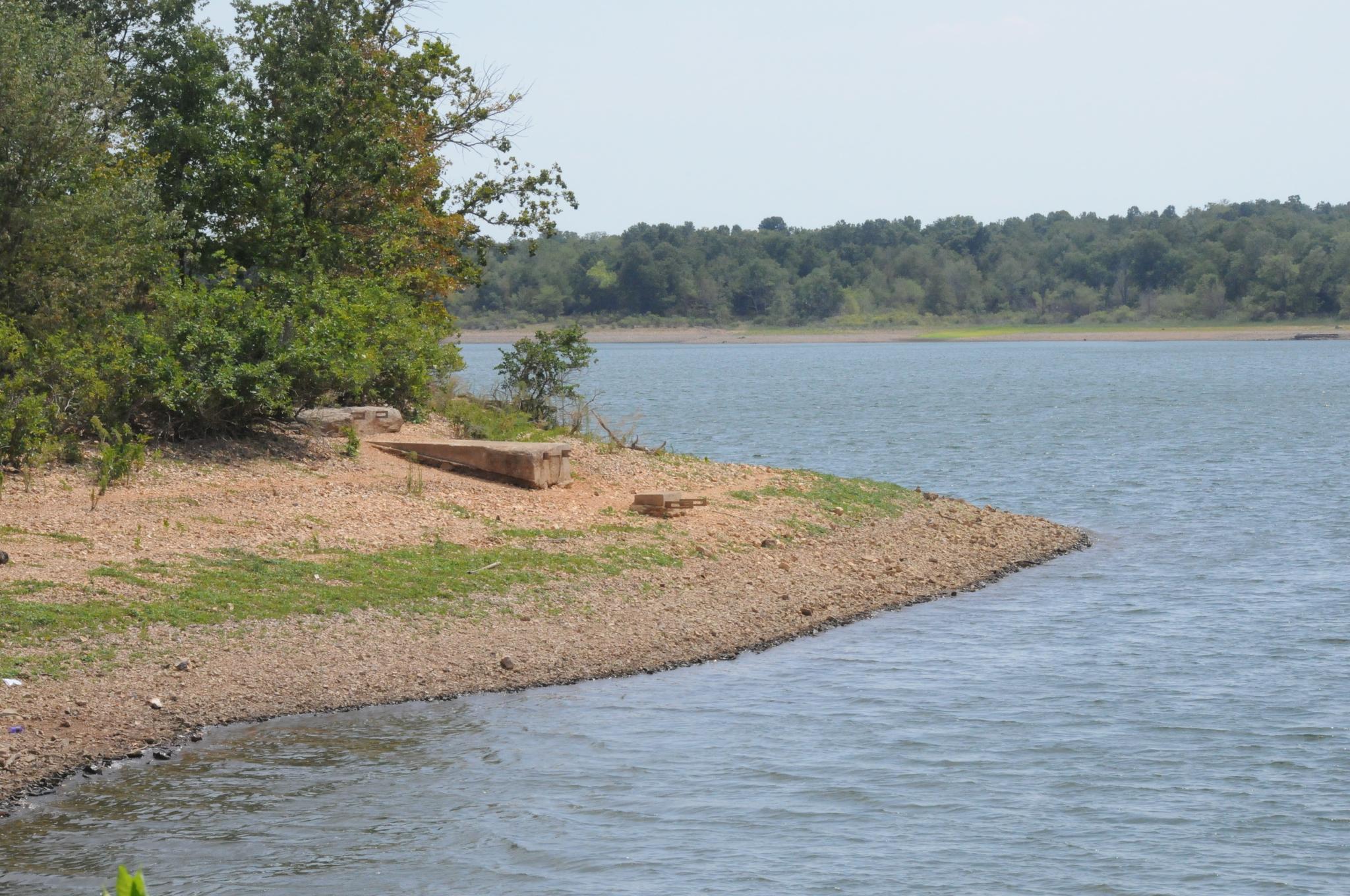 Bright day over a calm lake, rocky shore with trees and a wooden bench.