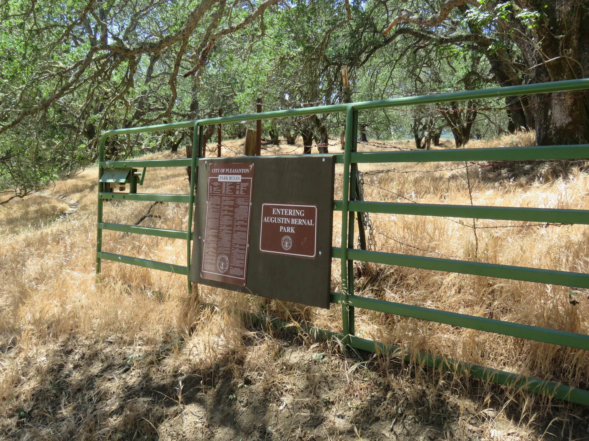 A green metal gate with signs in a field of dry grass and trees.