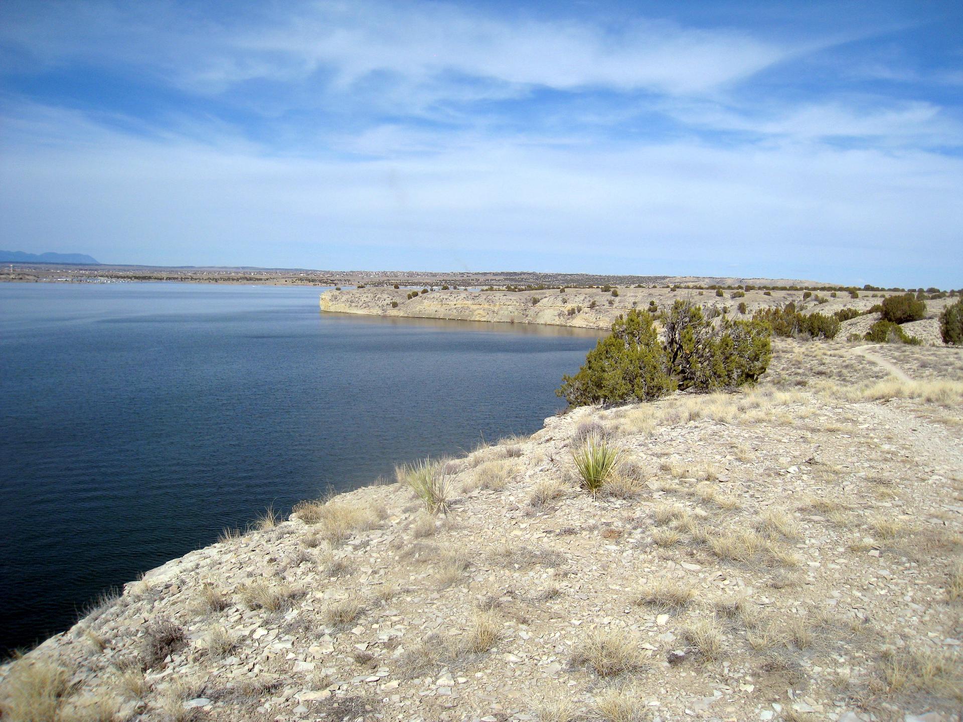Wide view of a calm lake bordered by arid, rocky cliffs under a bright sky.