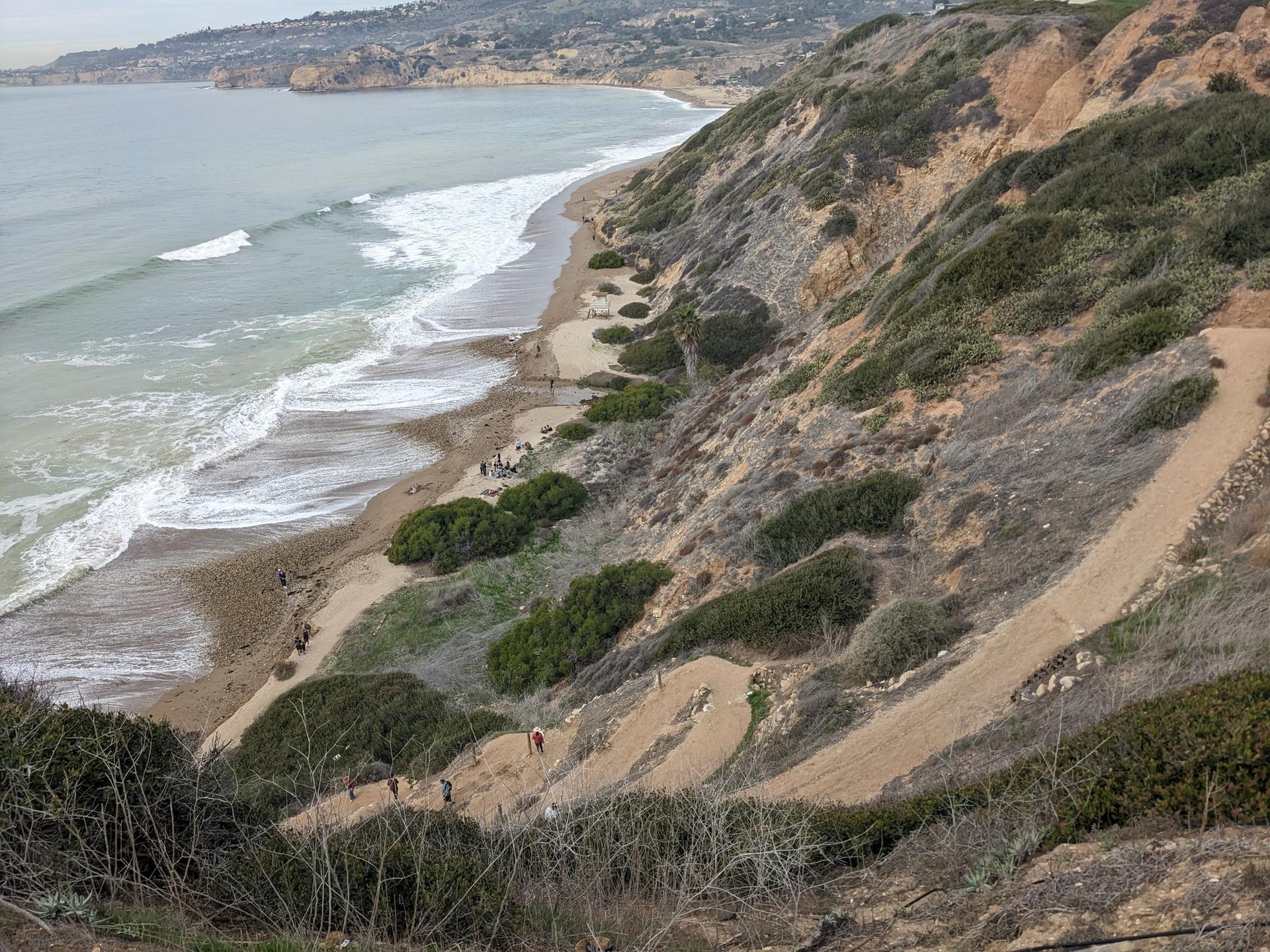 Overhead view of a steep coastal cliff, beach, ocean, and people walking.