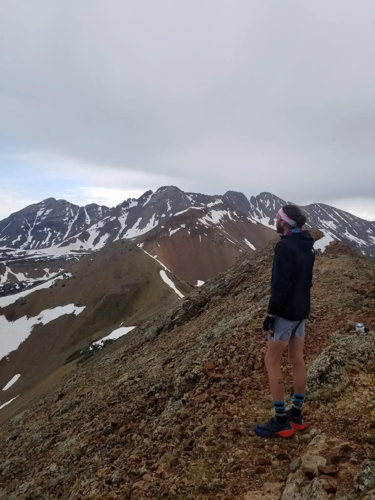 Person on a rocky mountain ridge overlooking snow-capped peaks under cloudy sky.