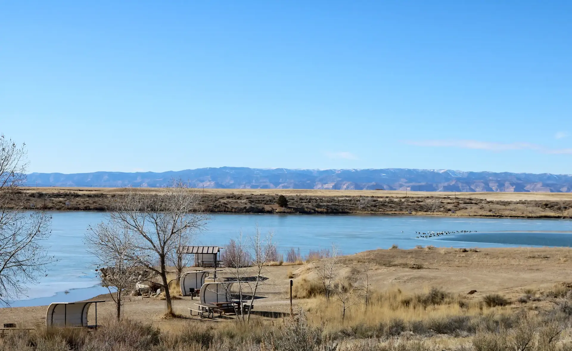 River, dry landscape with bare trees, shelters, and distant mountains on a sunny day.