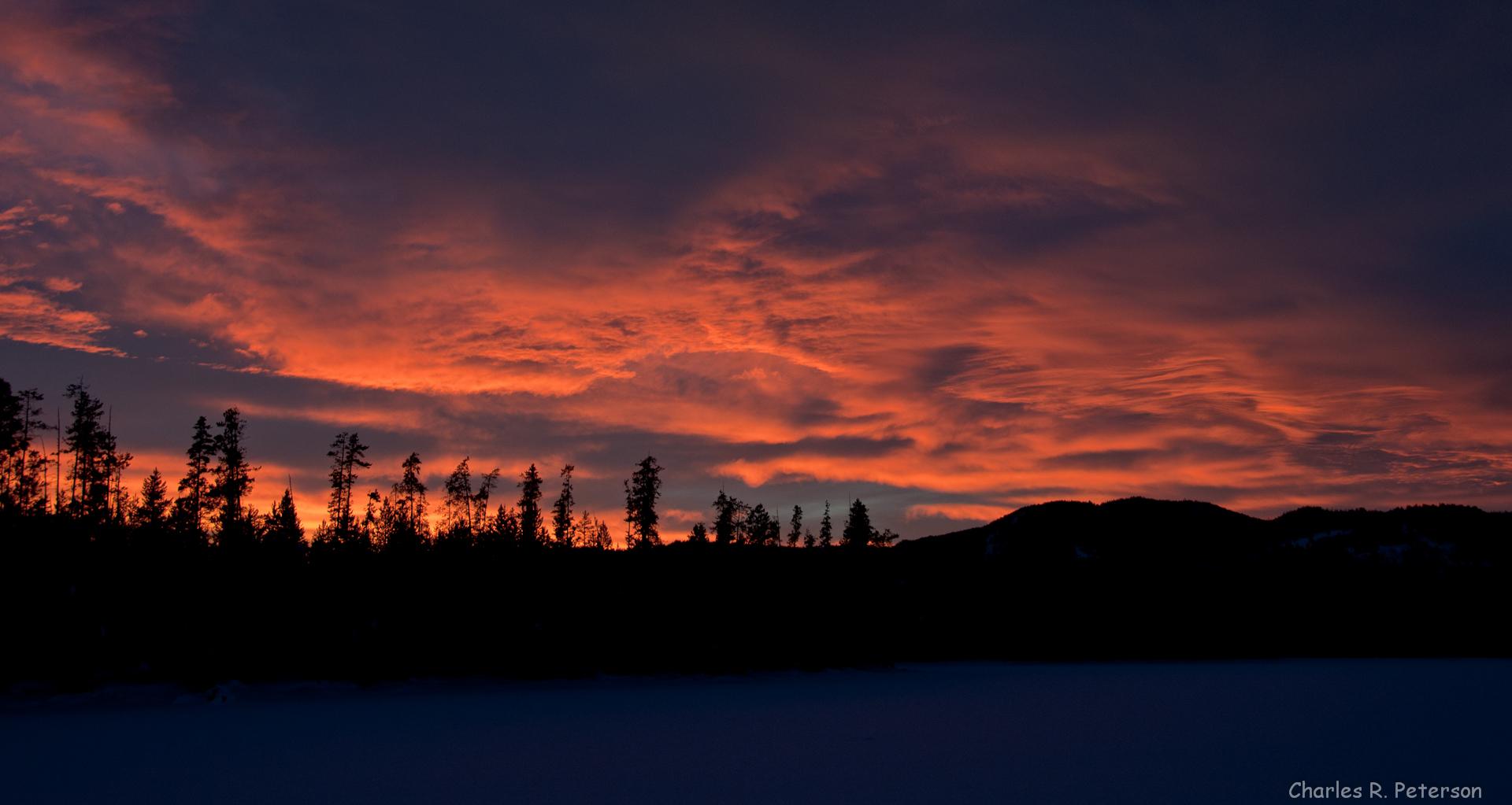 Vibrant orange and red sunset clouds over silhouetted trees and dark water.