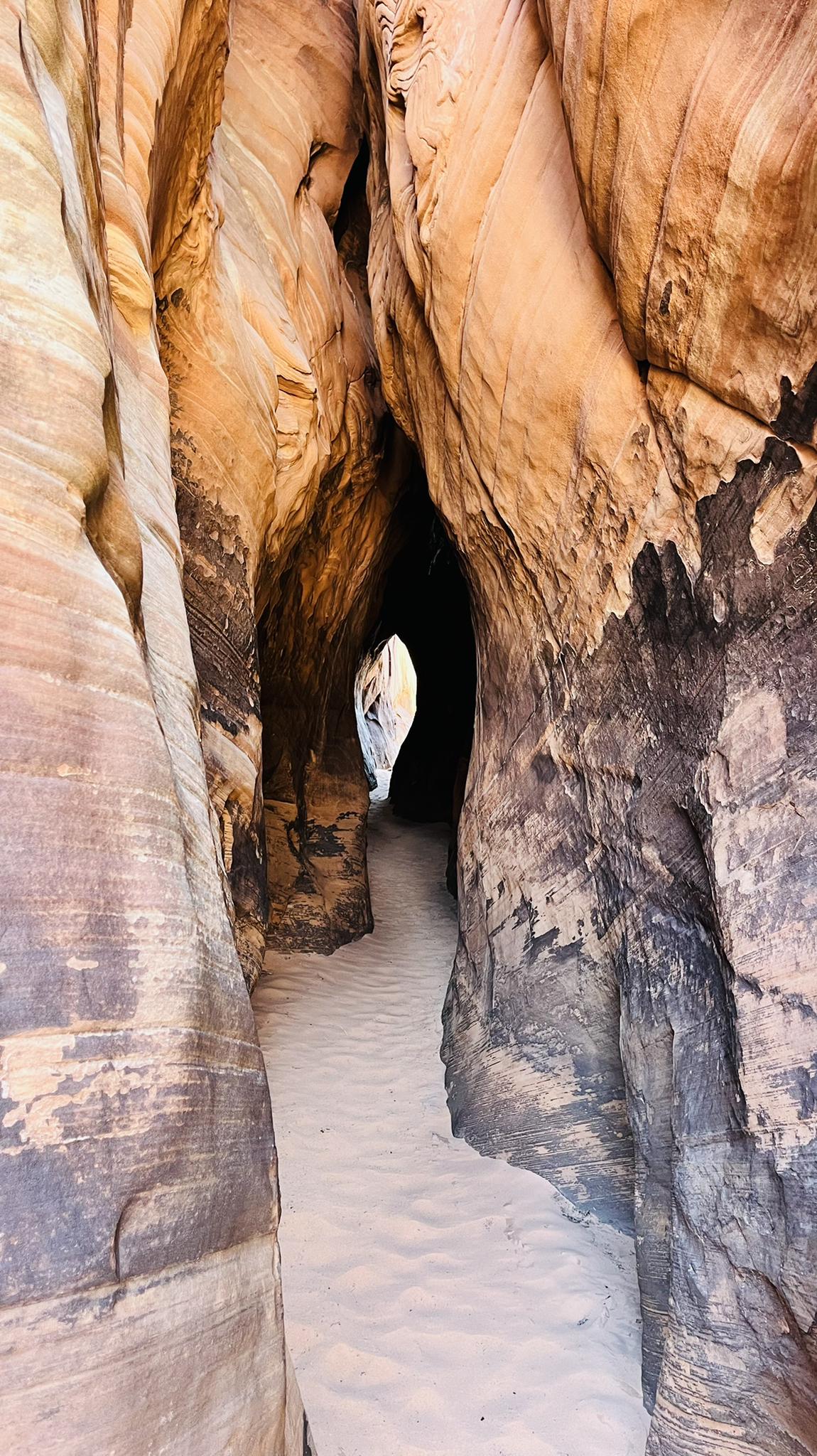 Narrow slot canyon with sandy floor and dark, light striped rock walls.