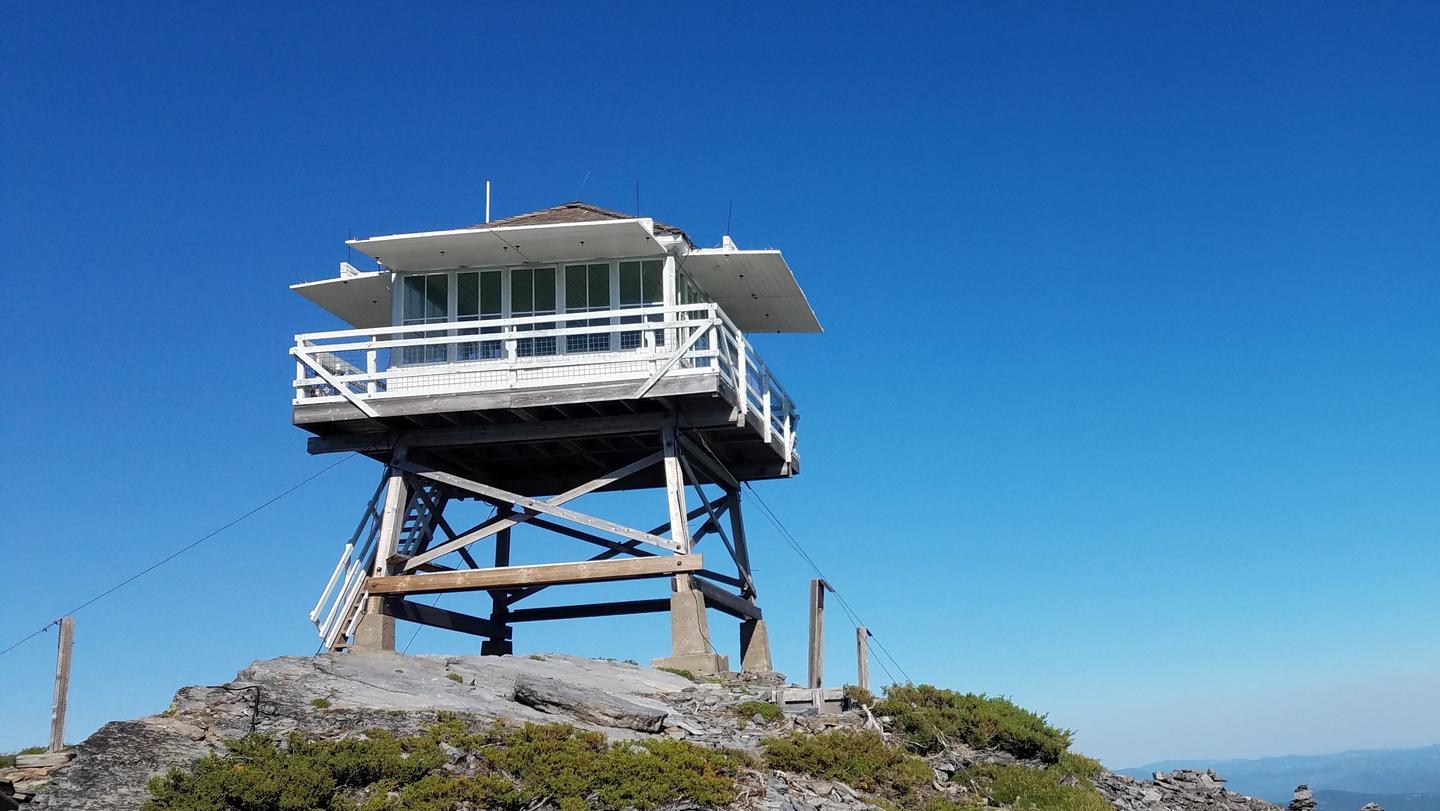 White fire lookout tower on a rocky peak under a clear blue sky.
