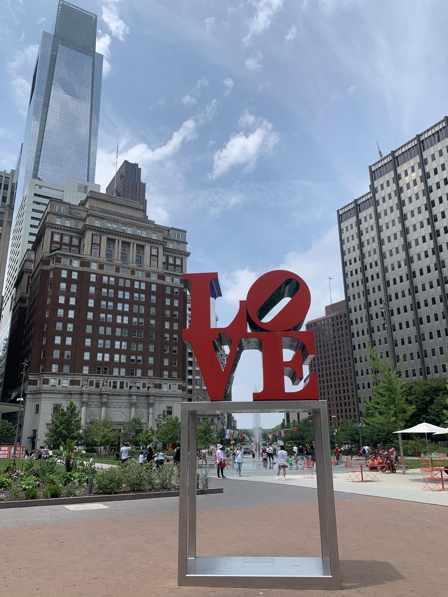 Red 'LOVE' sculpture in an urban park with tall buildings and people.