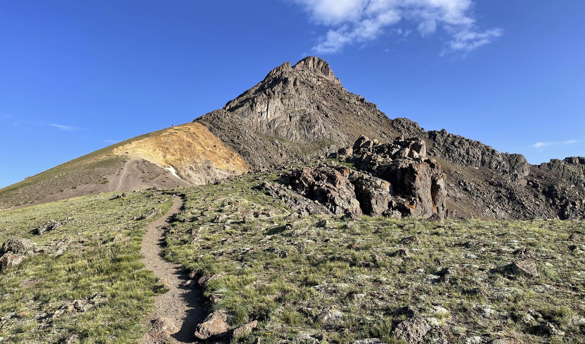 A winding dirt trail leads towards a large, rocky mountain peak.