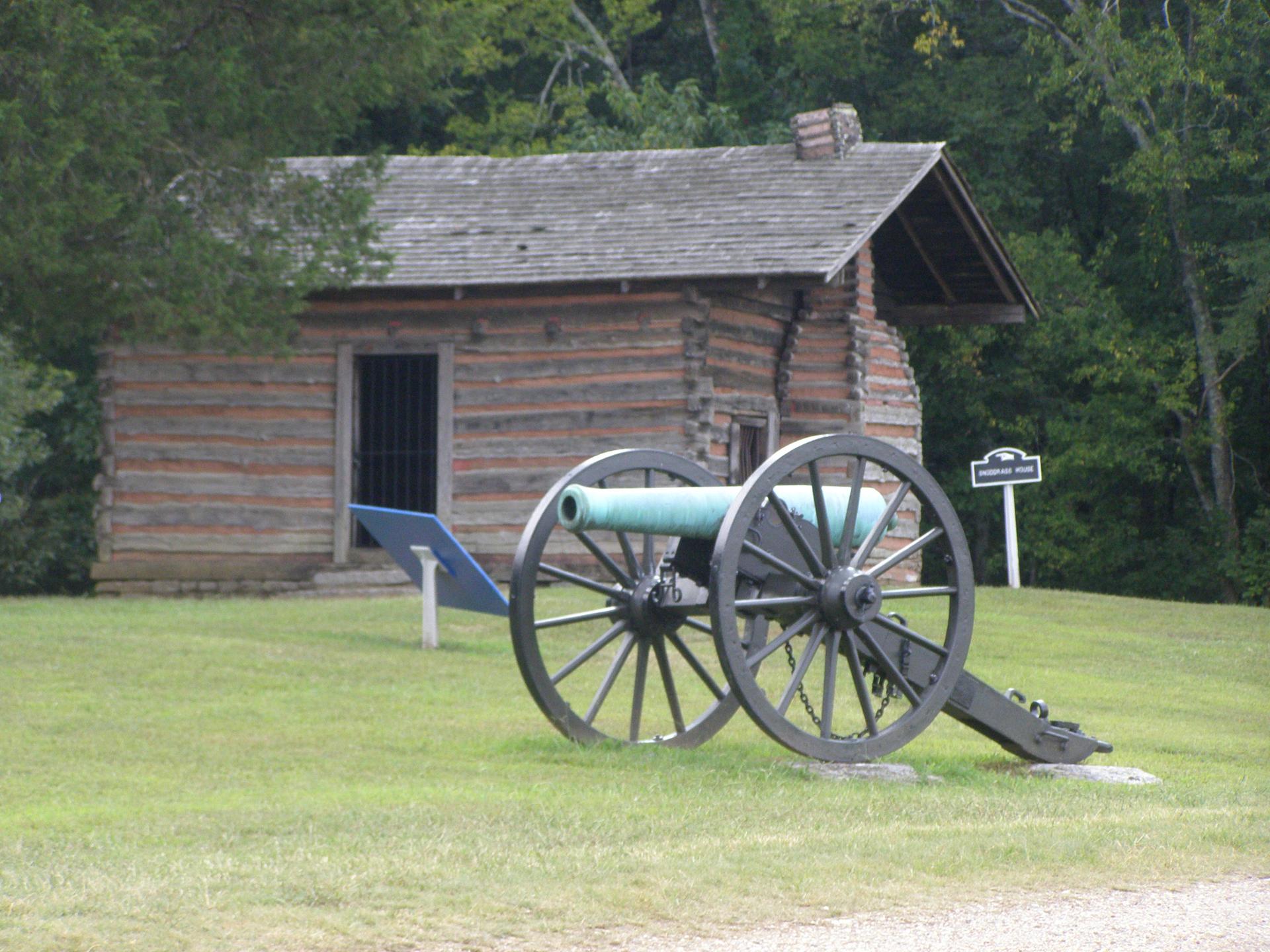 An old log cabin behind a cannon on a grassy hill, surrounded by trees.