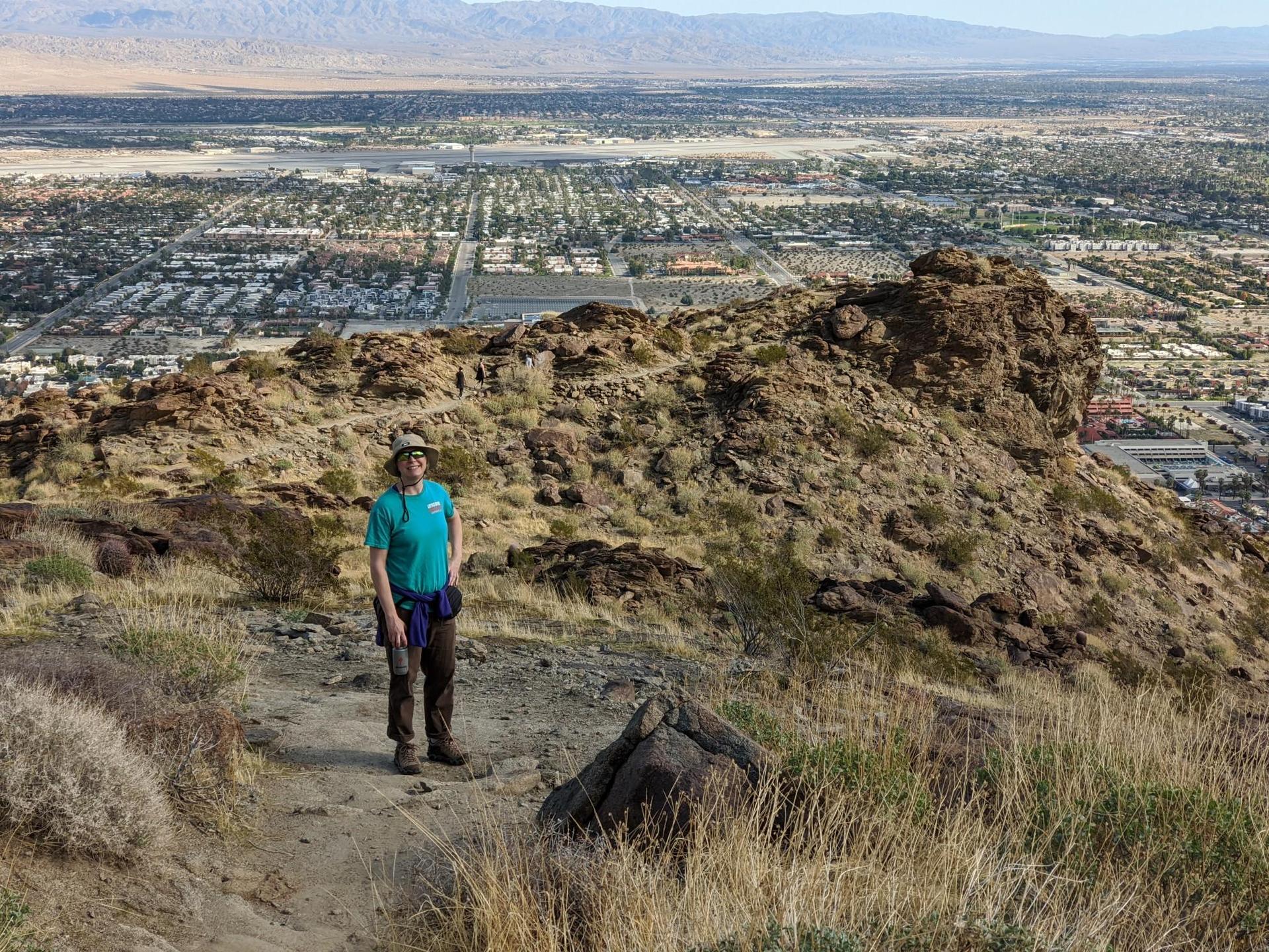 A person stands on a rocky trail overlooking a city on a sunny day.