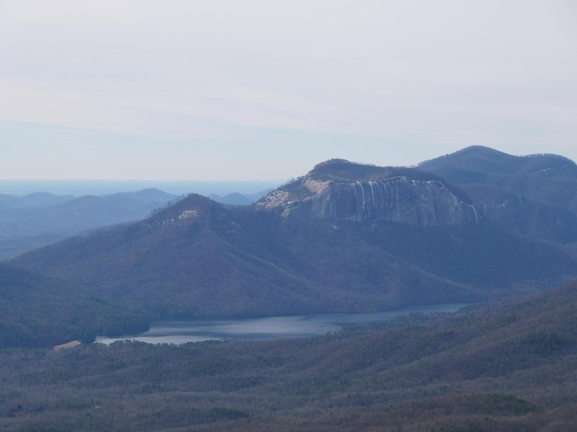Hazy view of a mountain range with a large rock face and a calm lake.