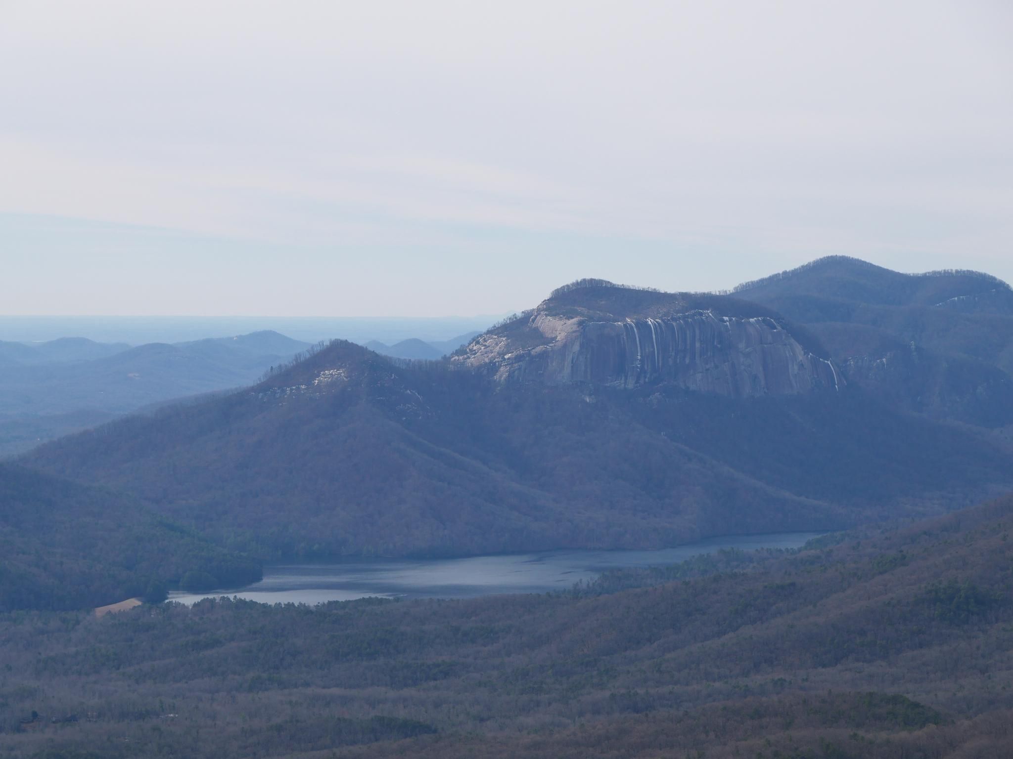 Hazy view of a mountain range with a large rock face and a calm lake. - Jones Gap loop