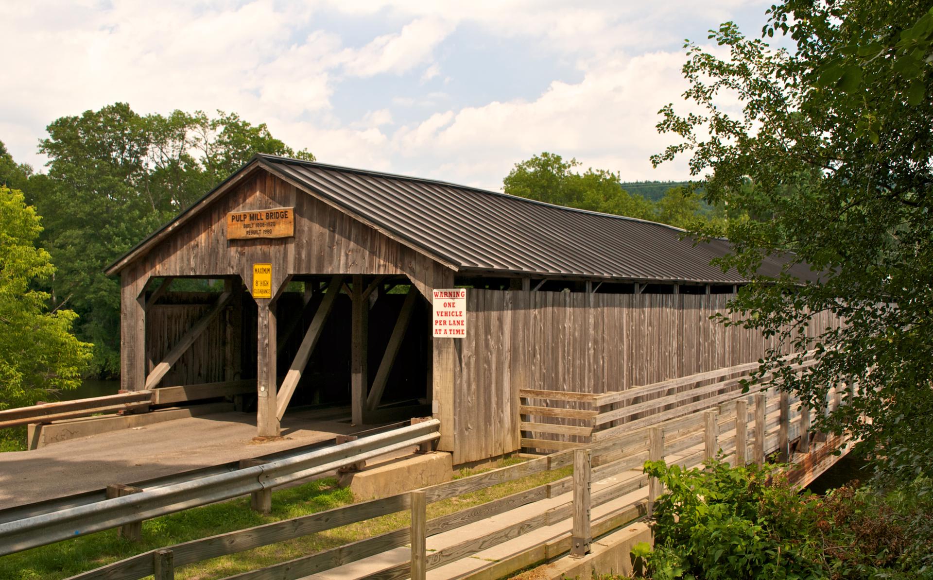 Rustic wooden covered bridge surrounded by green trees under a bright sky.