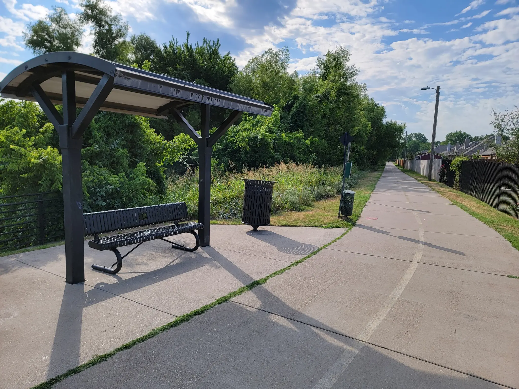 A covered bench and trash can next to a paved path on a sunny day.