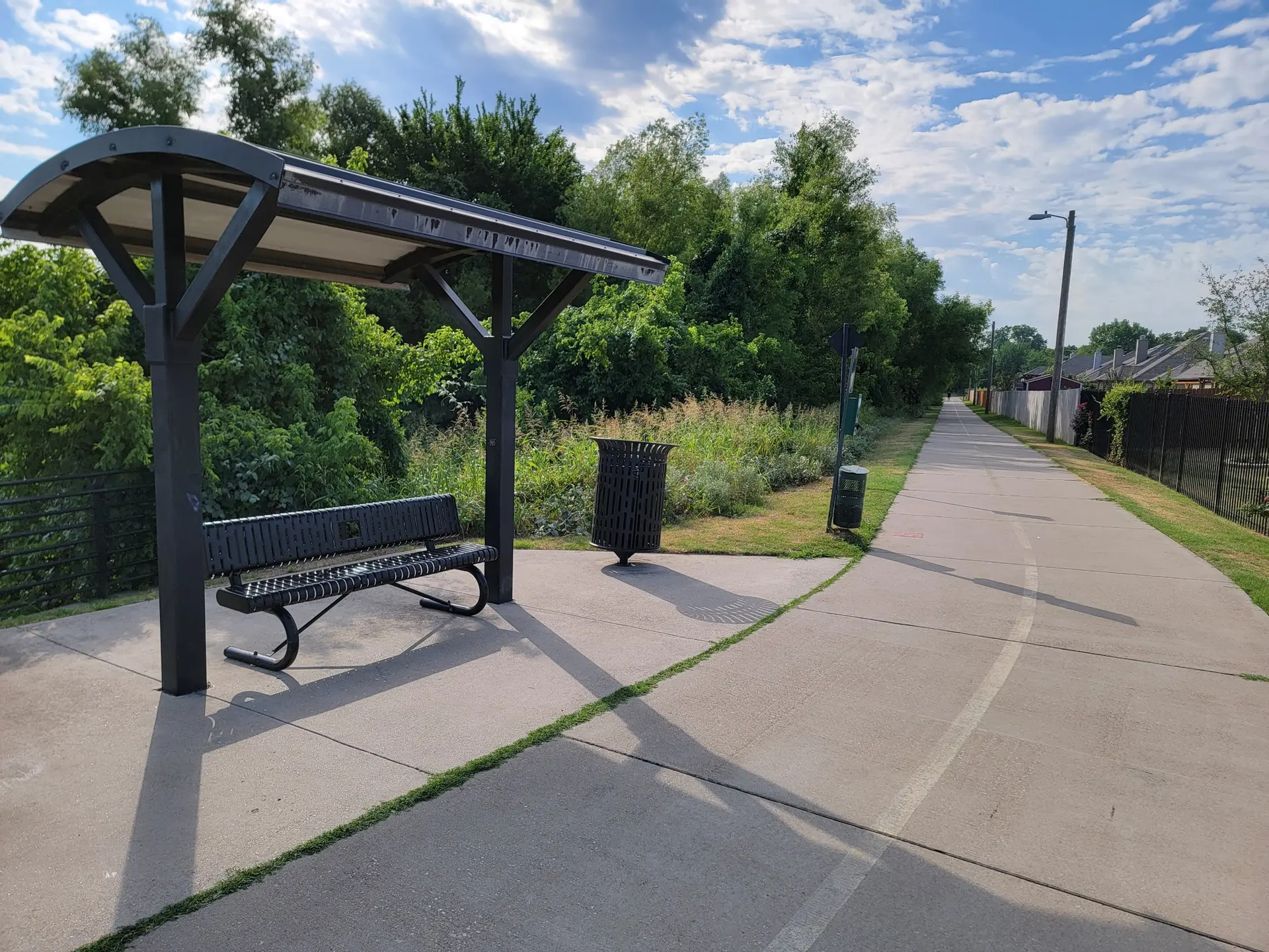 A covered bench and trash can next to a paved path on a sunny day.