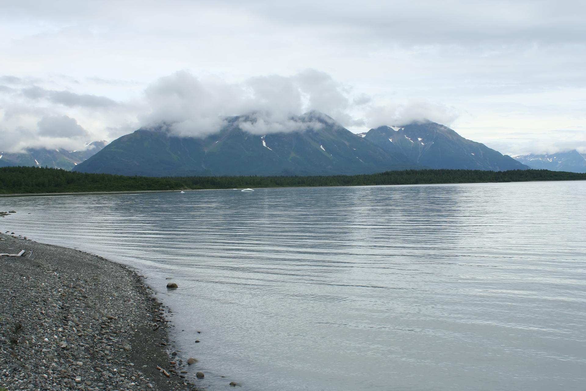 A calm lake with a rocky shore, backed by cloud-covered, snow-capped mountains.