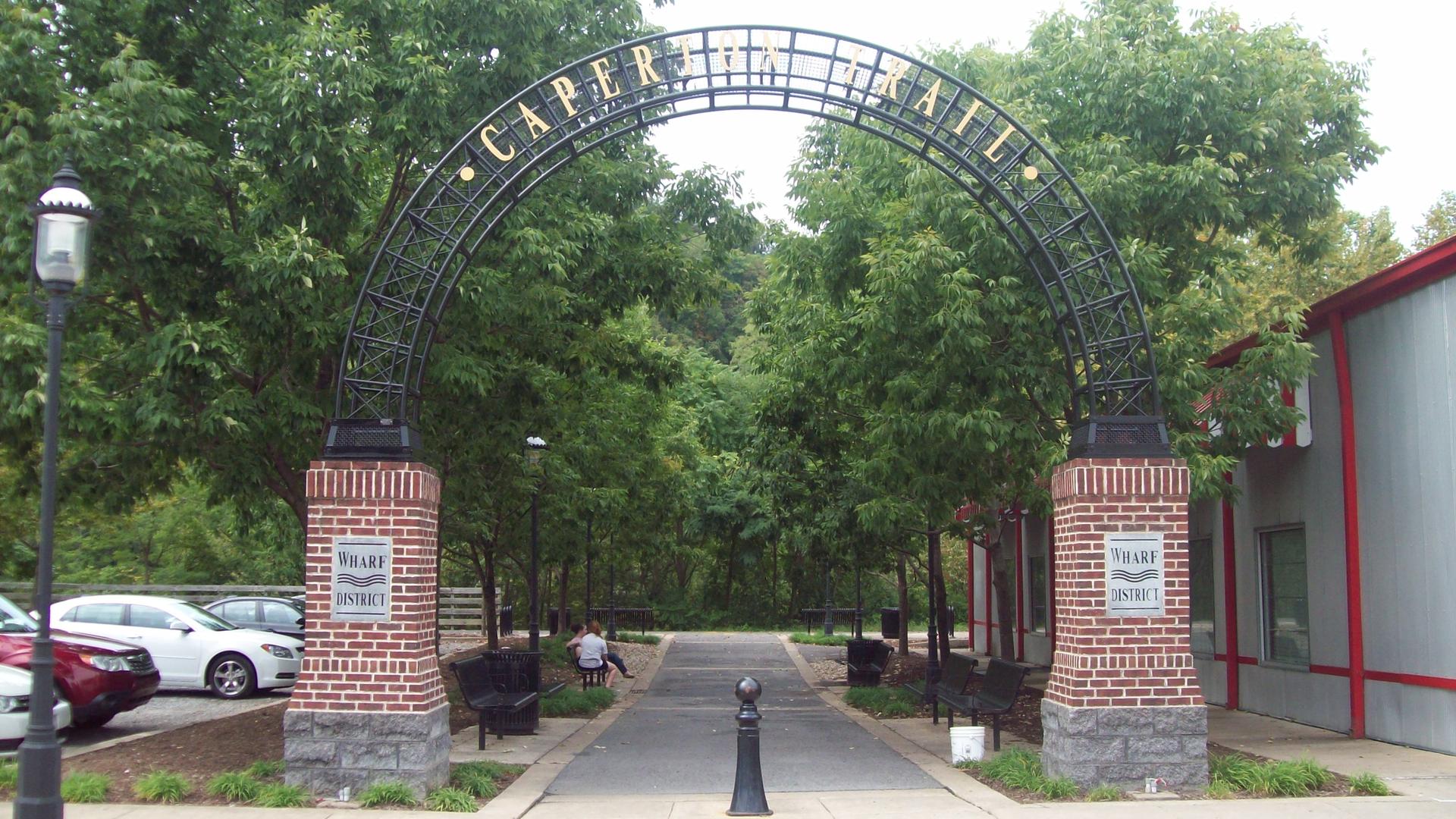 Large metal archway over a paved path, lush trees, and a person sitting.