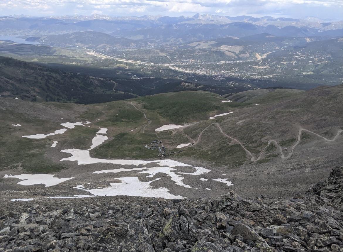 Rocky mountain landscape with snow patches and a distant town under an overcast sky.