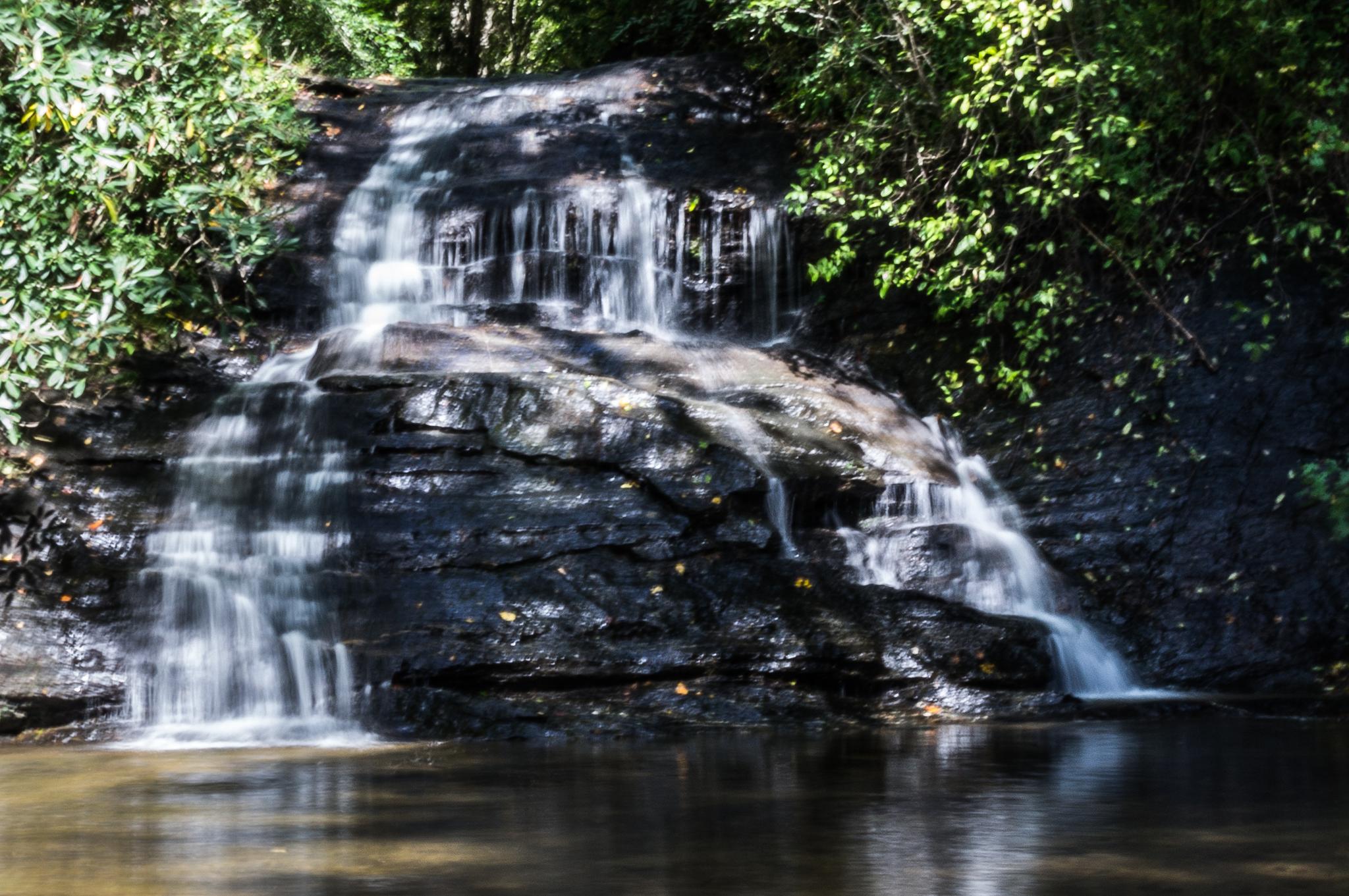 A multi-tiered waterfall cascades over dark rocks into a pool, surrounded by green foliage.