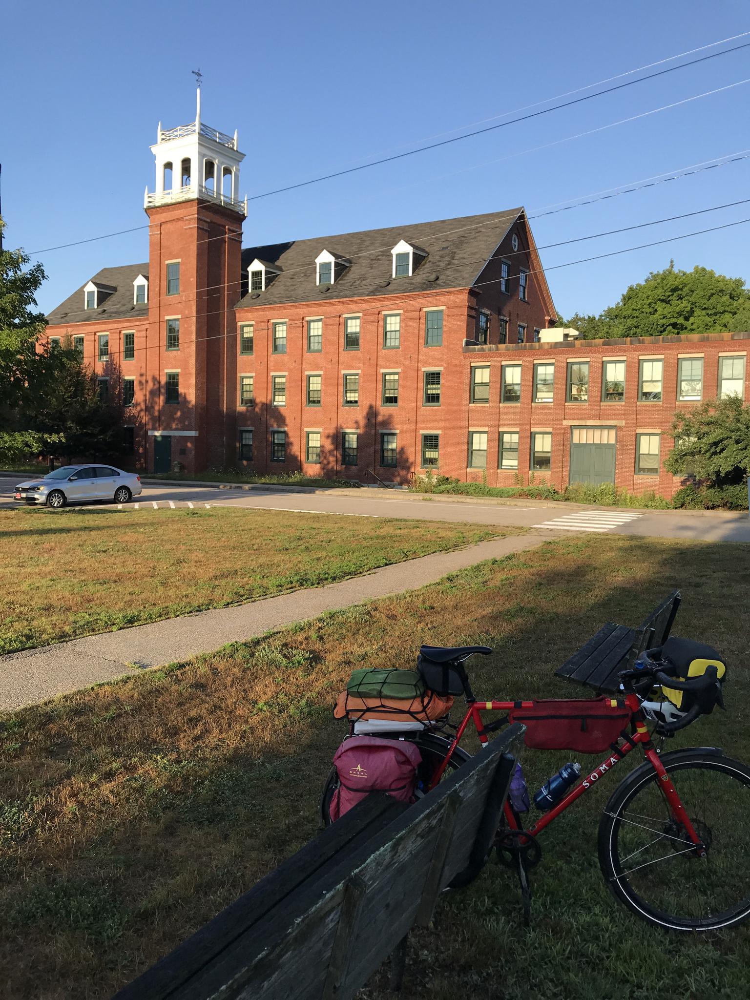 A packed touring bicycle rests in grass before a large brick building.