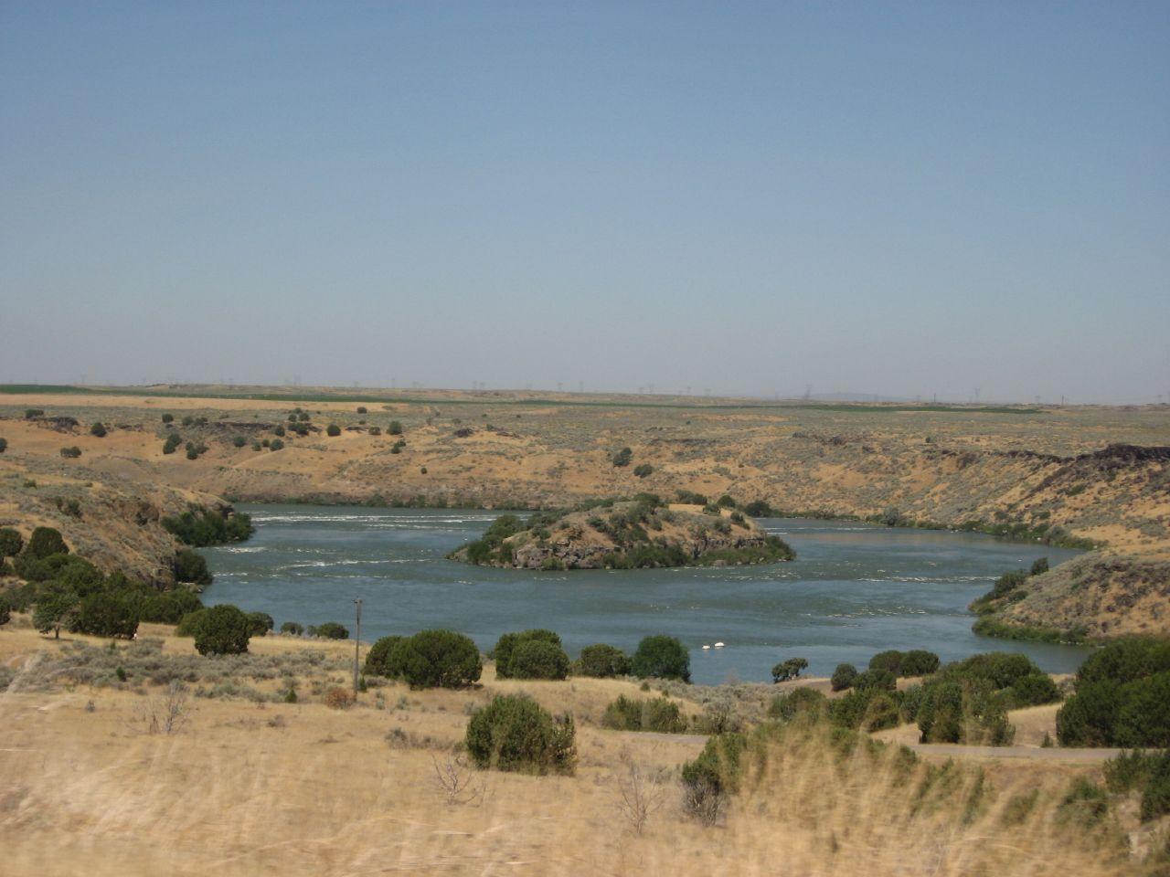 Wide river with an island, surrounded by dry, rocky hills under a clear sky.