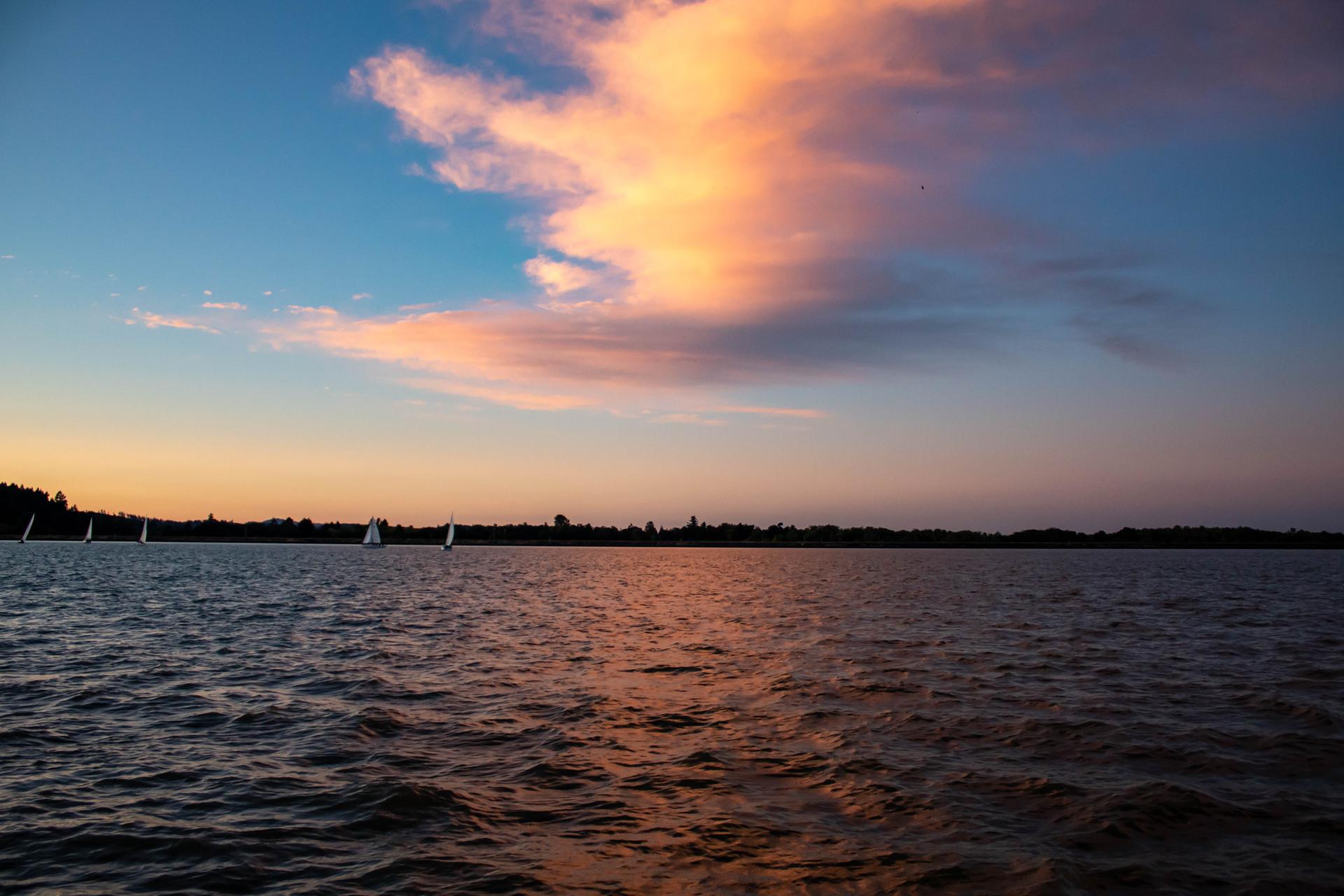 Serene sunset over water with sailboats and vibrant clouds.