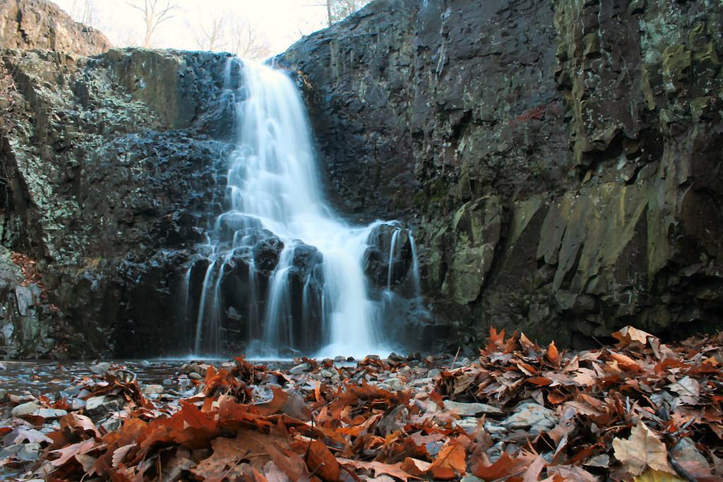 Cascading waterfall over dark rocks, surrounded by fallen autumn leaves.
