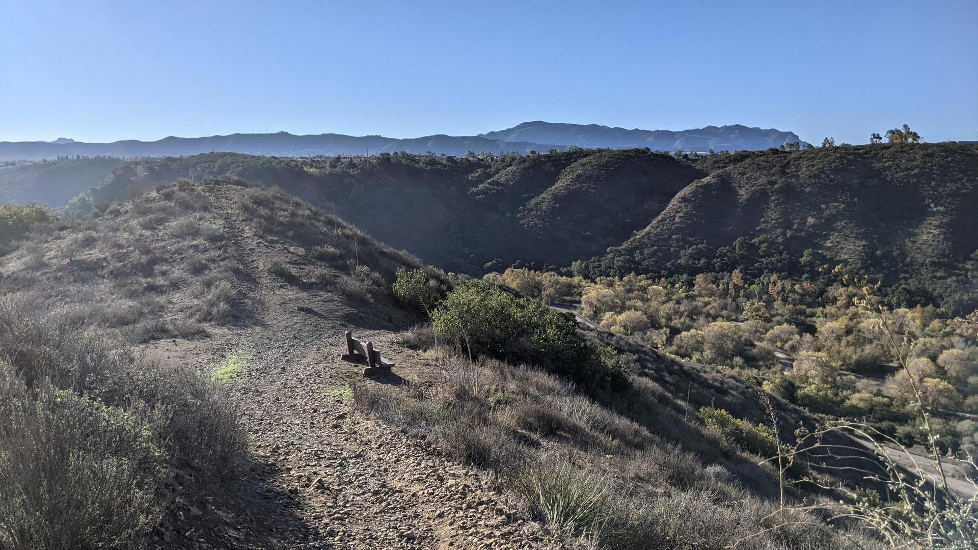 Two people rest on a sunny, dry hillside trail overlooking valleys and mountains.