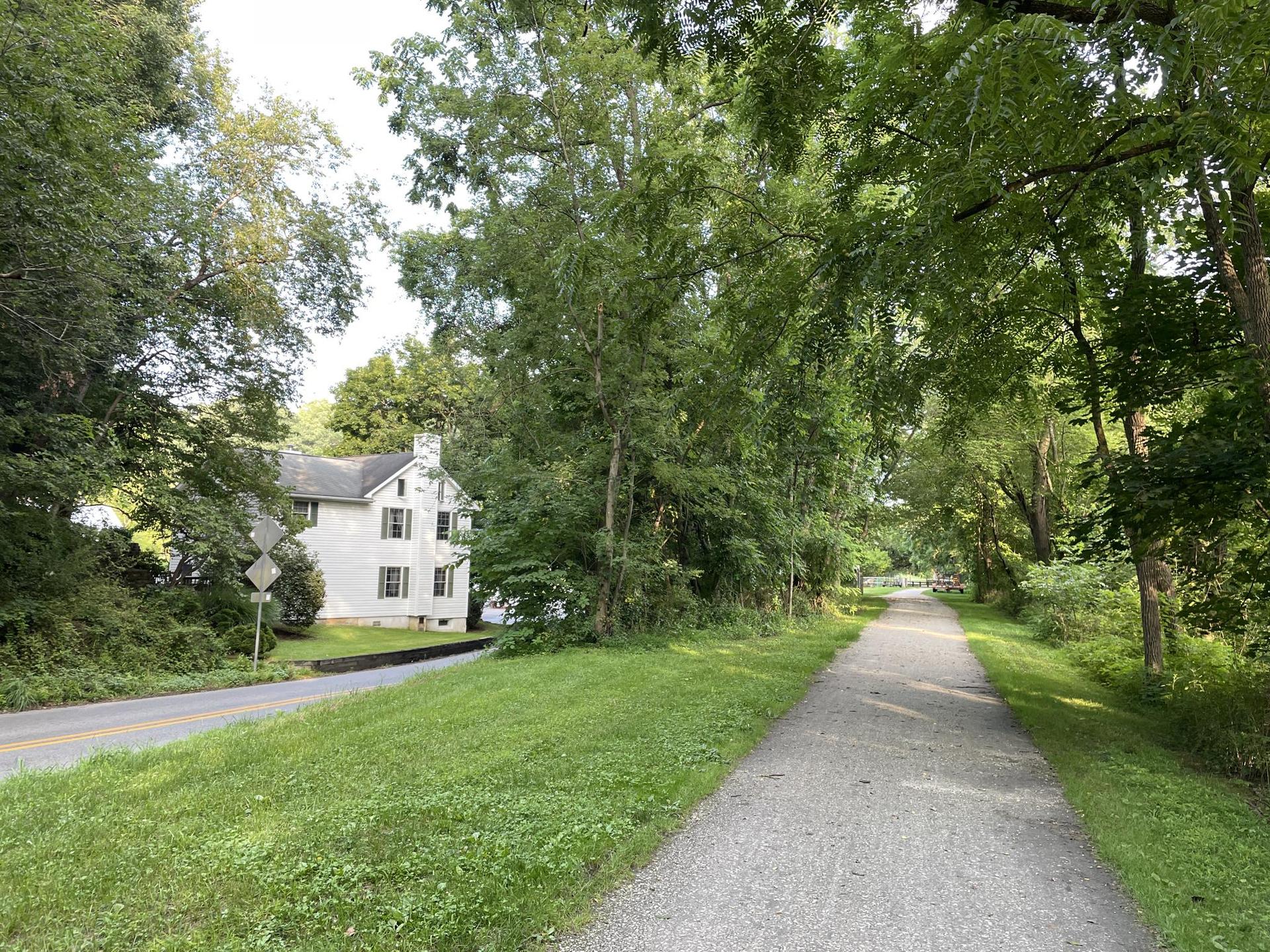 A gravel path runs beside a road, with a white house and green trees.