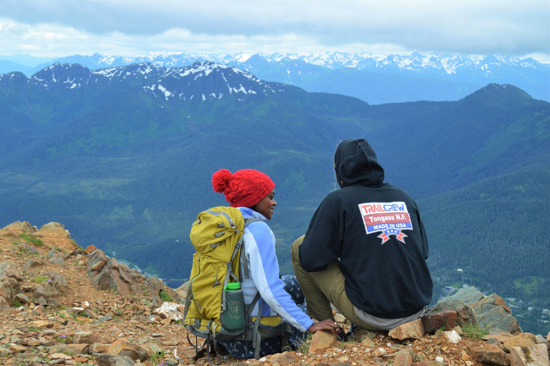 Two hikers sit on a mountain peak, overlooking vast snowy ranges.