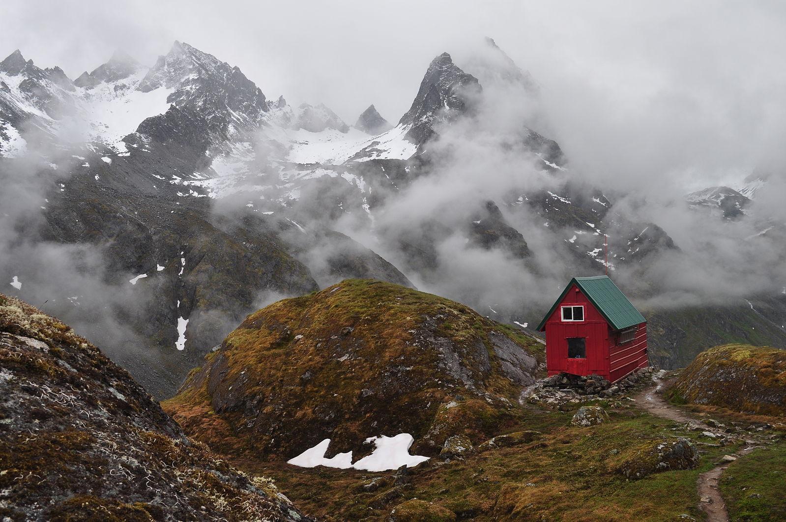 Red cabin on a rocky mountain slope with misty, snow-capped peaks.