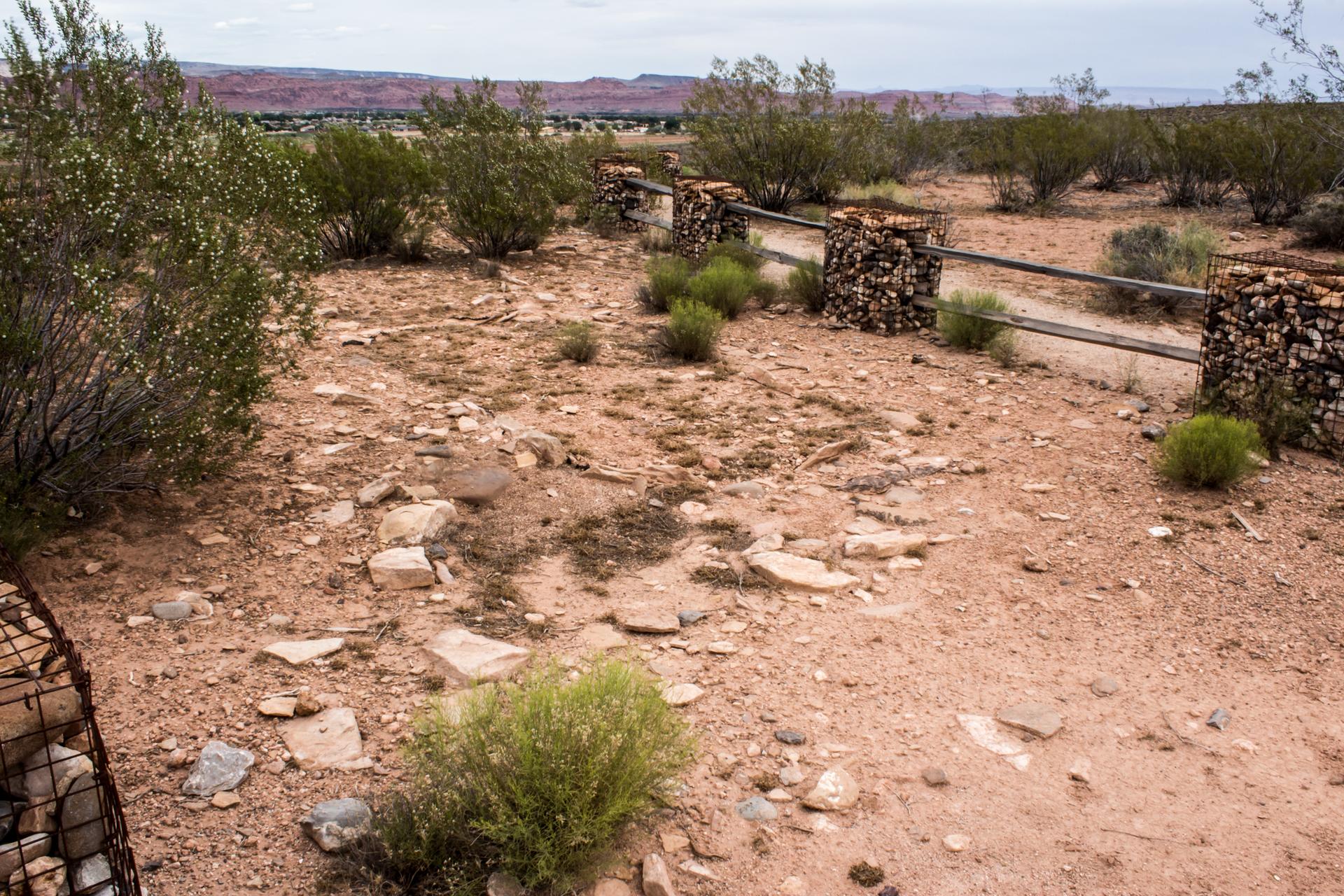 Gabion fence in a dry, rocky landscape with sparse bushes under an overcast sky.