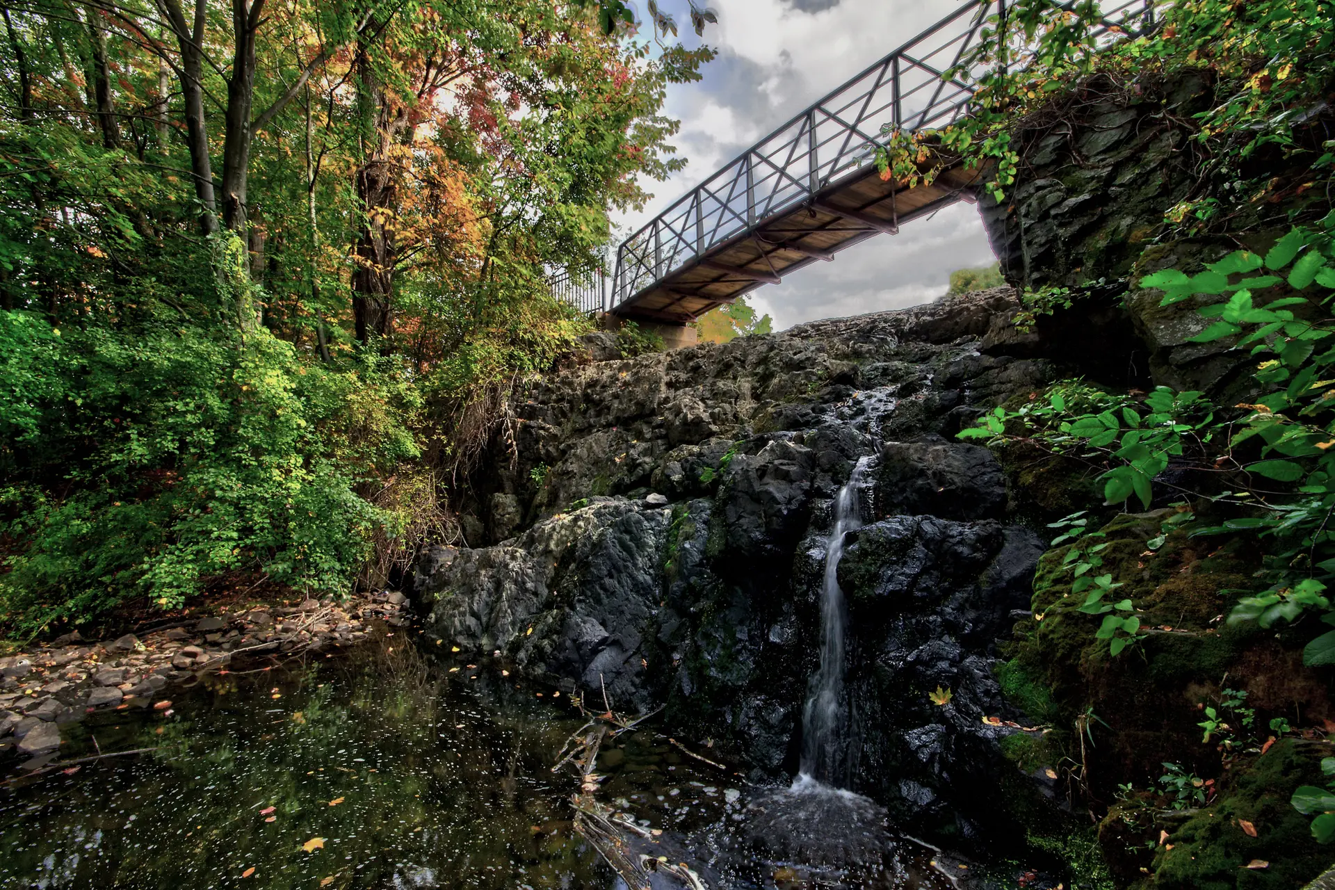 Waterfall cascades over dark rocks beneath a metal bridge, with vibrant foliage.