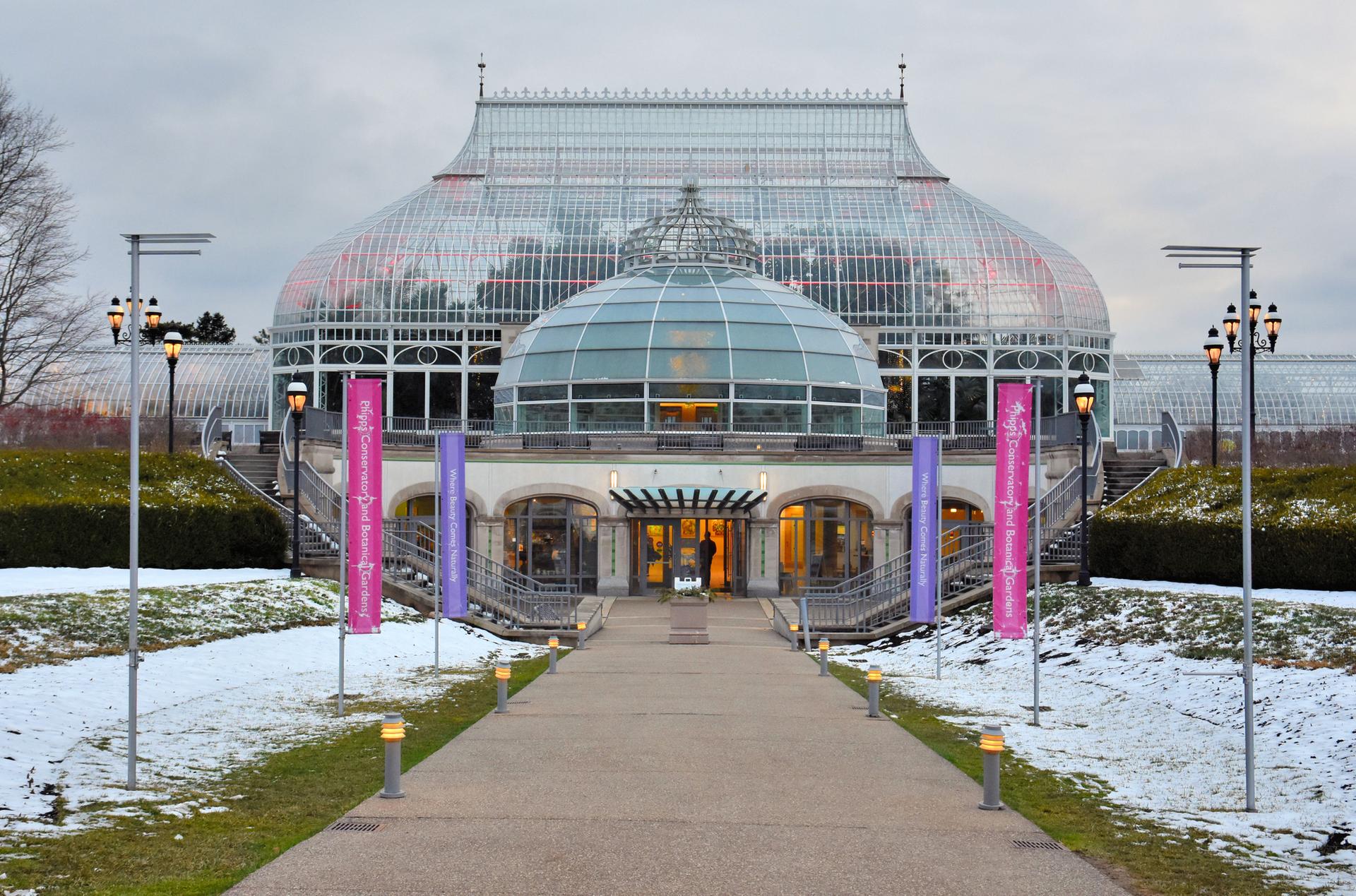 Grand glass conservatory building with snow on the ground under an overcast sky.